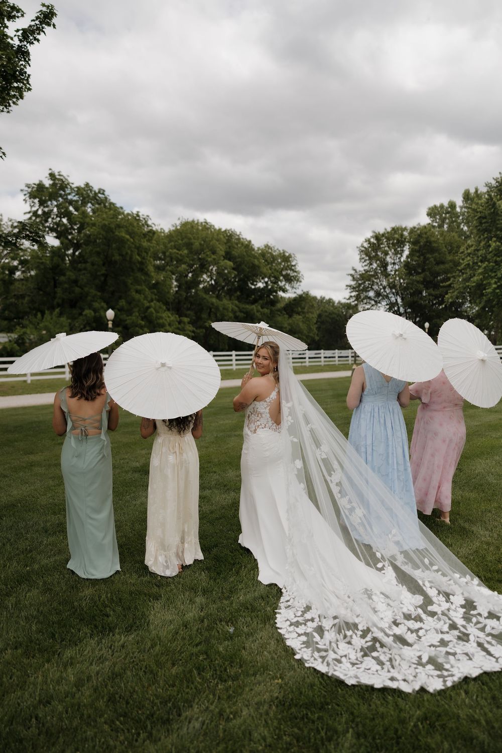 Bride with cathedral veil poses with three bridesmaids holding white parasols on a green lawn outdoors.