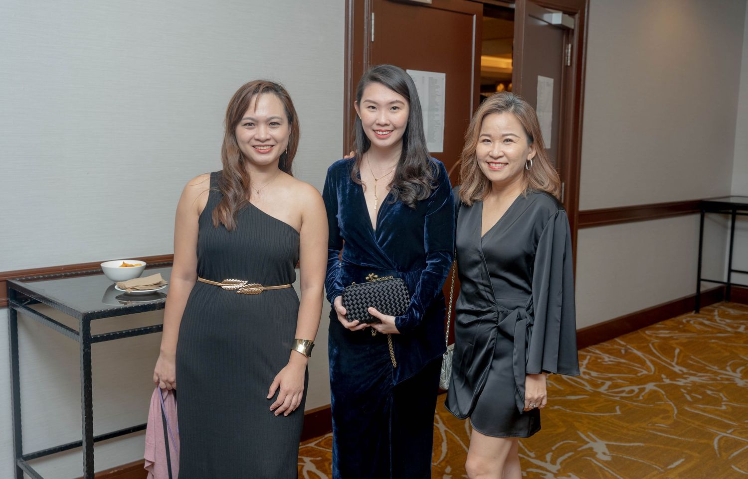 Three elegantly dressed attendees pose together at a formal evening event inside a hotel hallway.