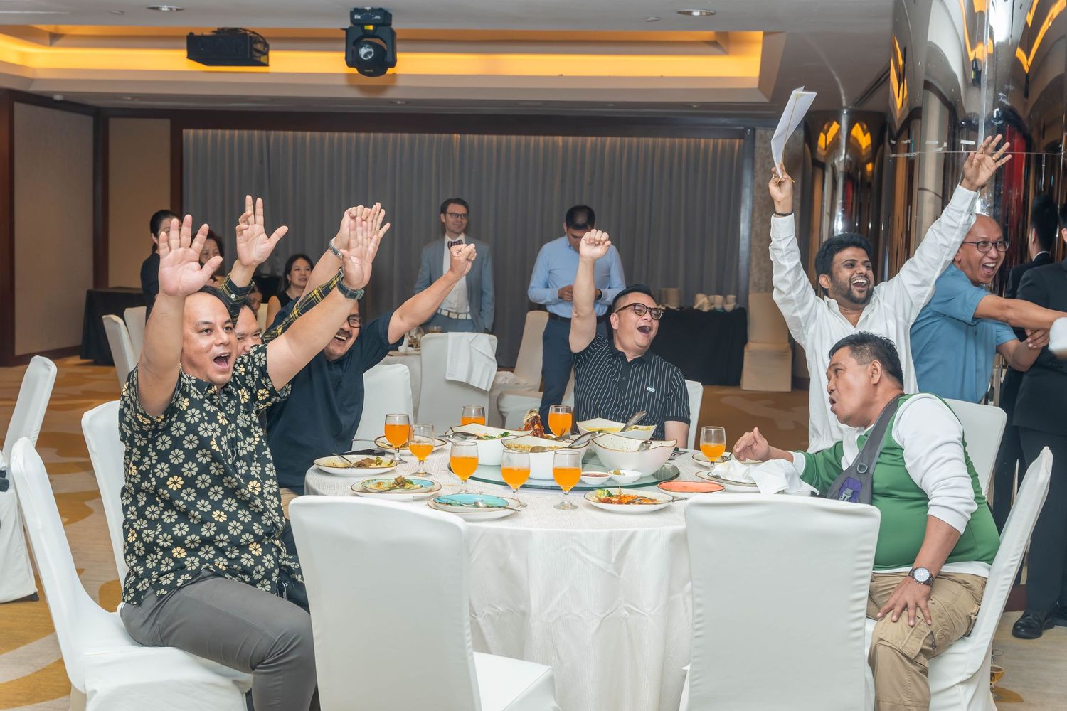Enthusiastic dinner guests raising arms in celebration around decorated banquet table.