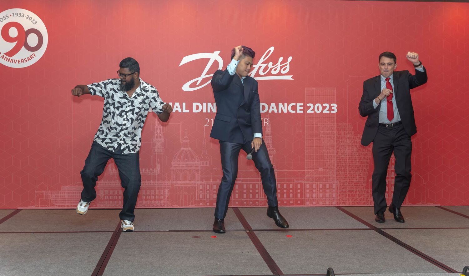 Three people dancing and posing energetically against a red backdrop with Danfoss branding.