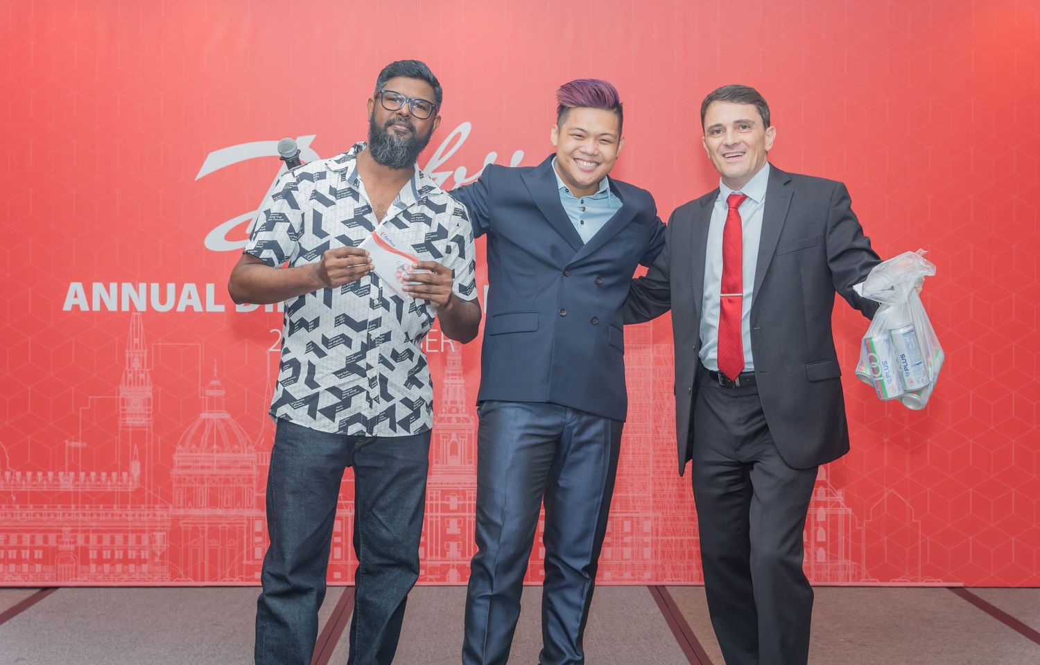 Three people striking fun poses together at a Danfoss annual event against a red background.