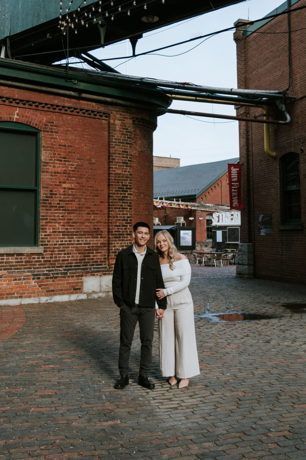 A couple standing on cobblestone street surrounded by historic brick buildings.