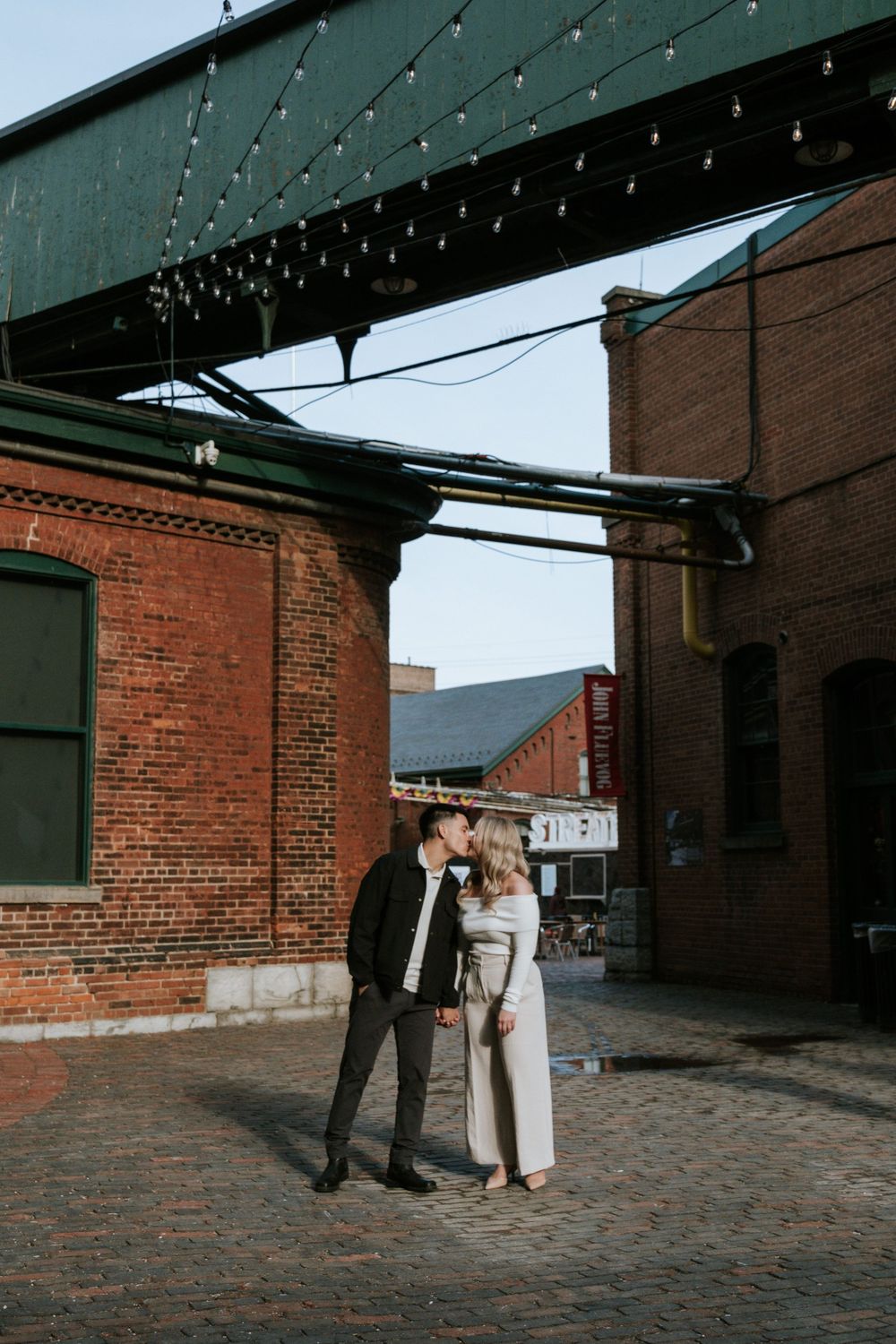 A romantic portrait beneath string lights against industrial brick architecture.