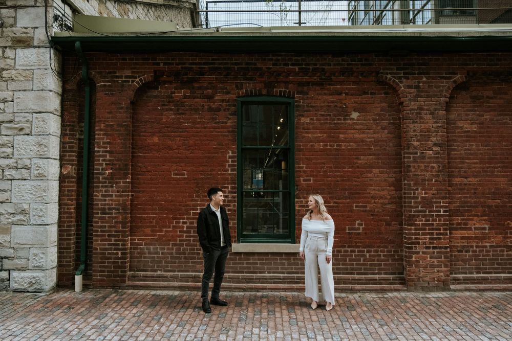 Couple in formal wear standing against rustic brick wall backdrop.