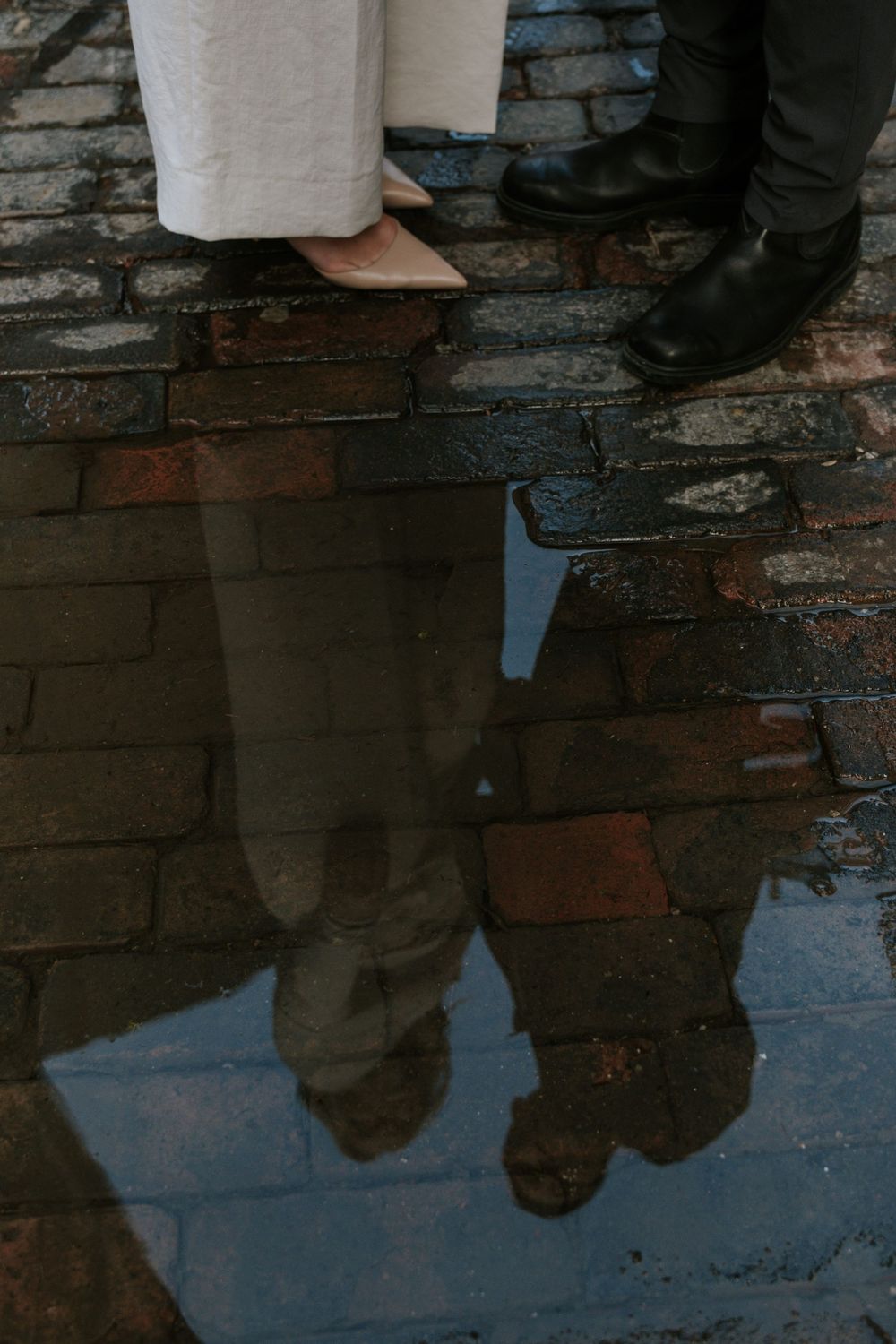 Reflection of a couple's feet in a puddle on brick pavement.