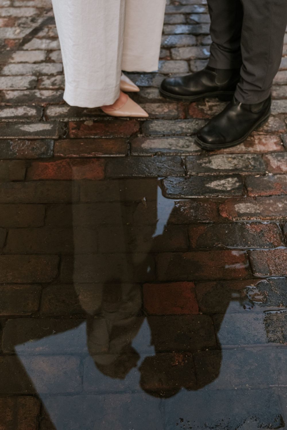 A romantic reflection of two pairs of feet standing on wet cobblestones, one in black boots and one in light colored flats.