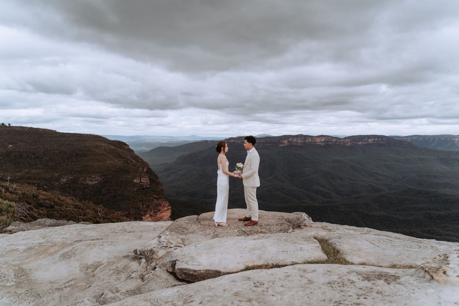 Couple in white outfits stands on rocky mountain cliff edge under dramatic cloudy sky.