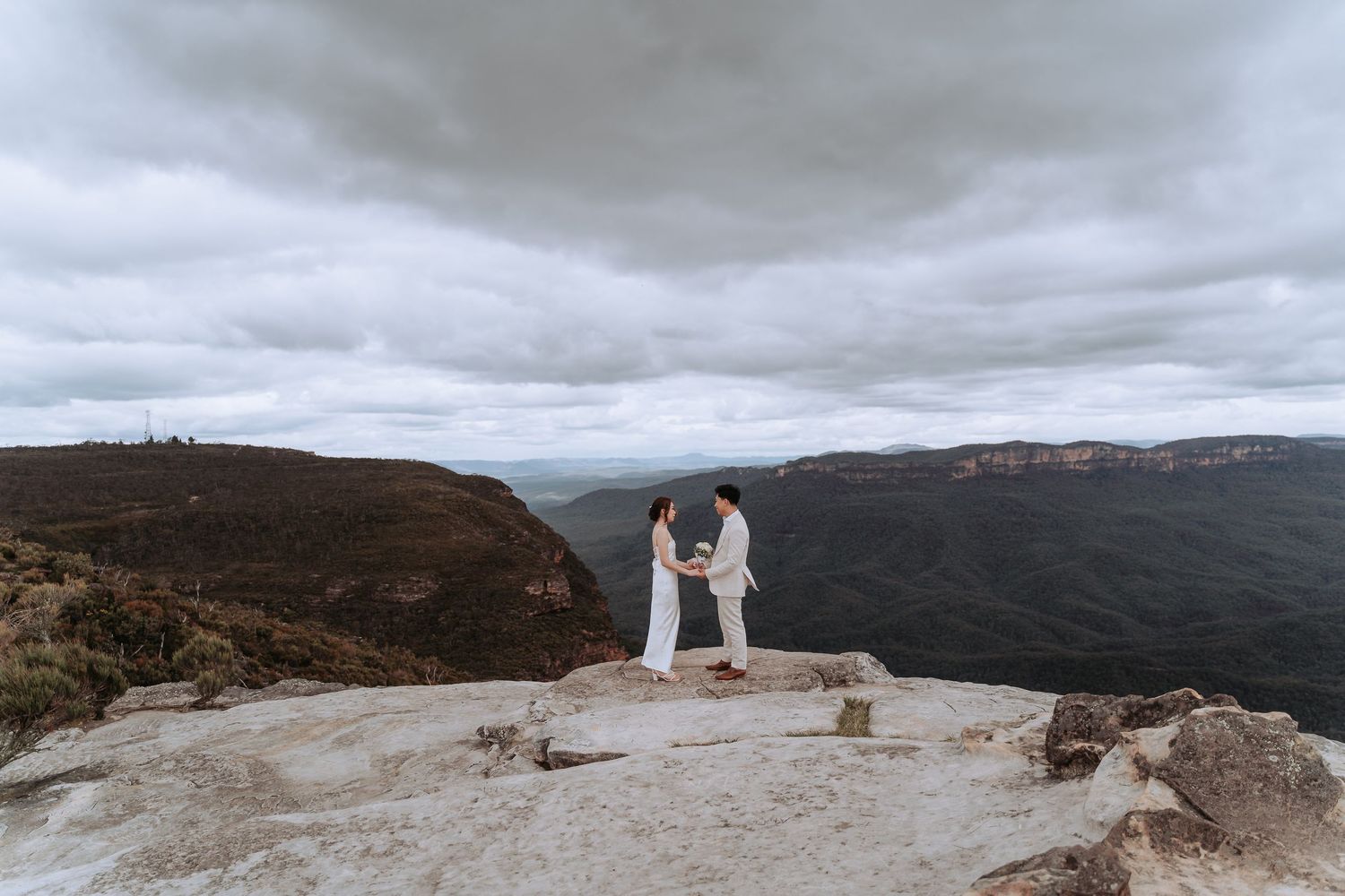 Two people in white clothing embrace on mountain lookout with cloudy valley vista.