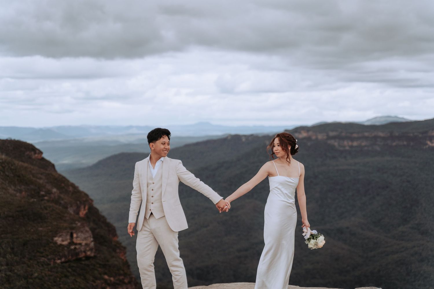 Couple in elegant attire holding hands on mountain cliff with valley views.
