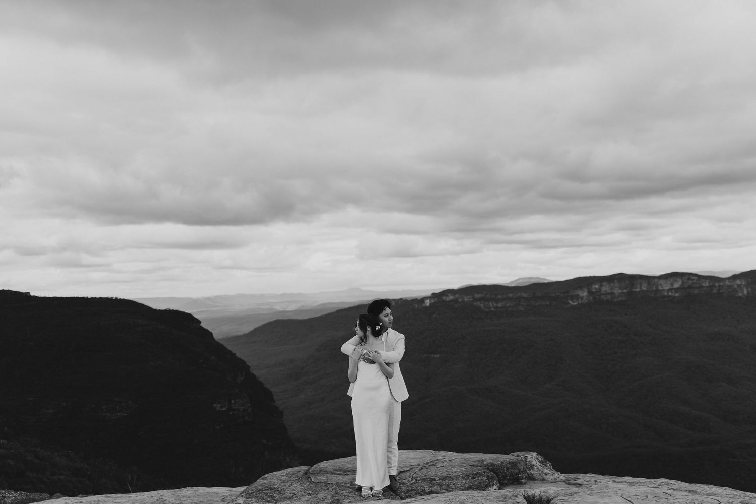 Romantic black and white photo of couple embracing on mountainous overlook.