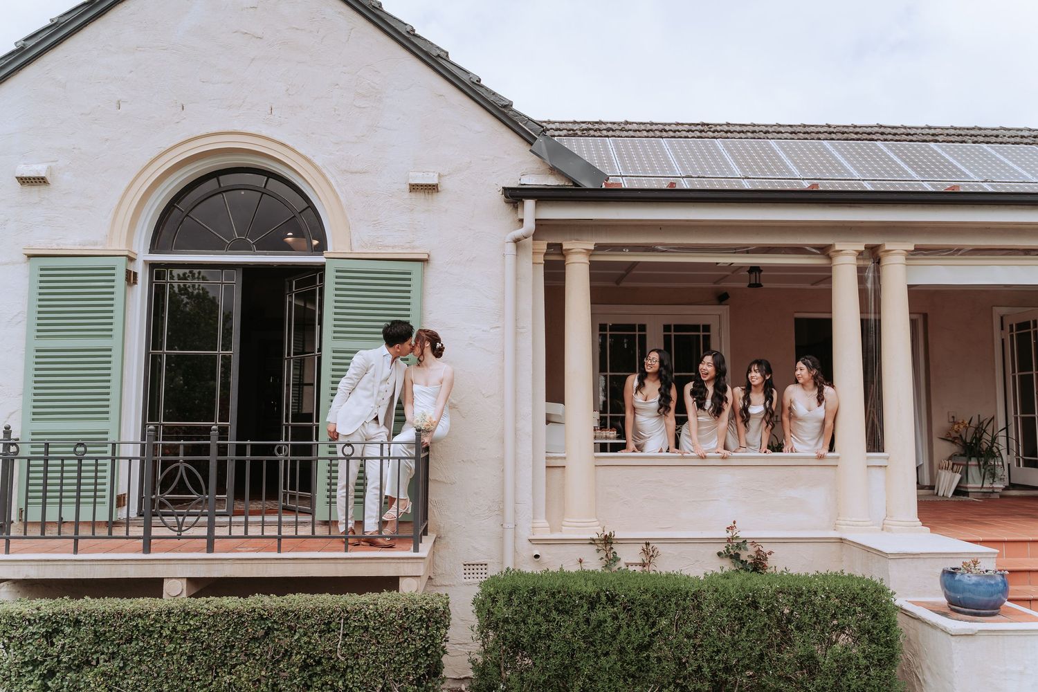Elegant couple on balcony of classic white colonial-style house with green shutters.