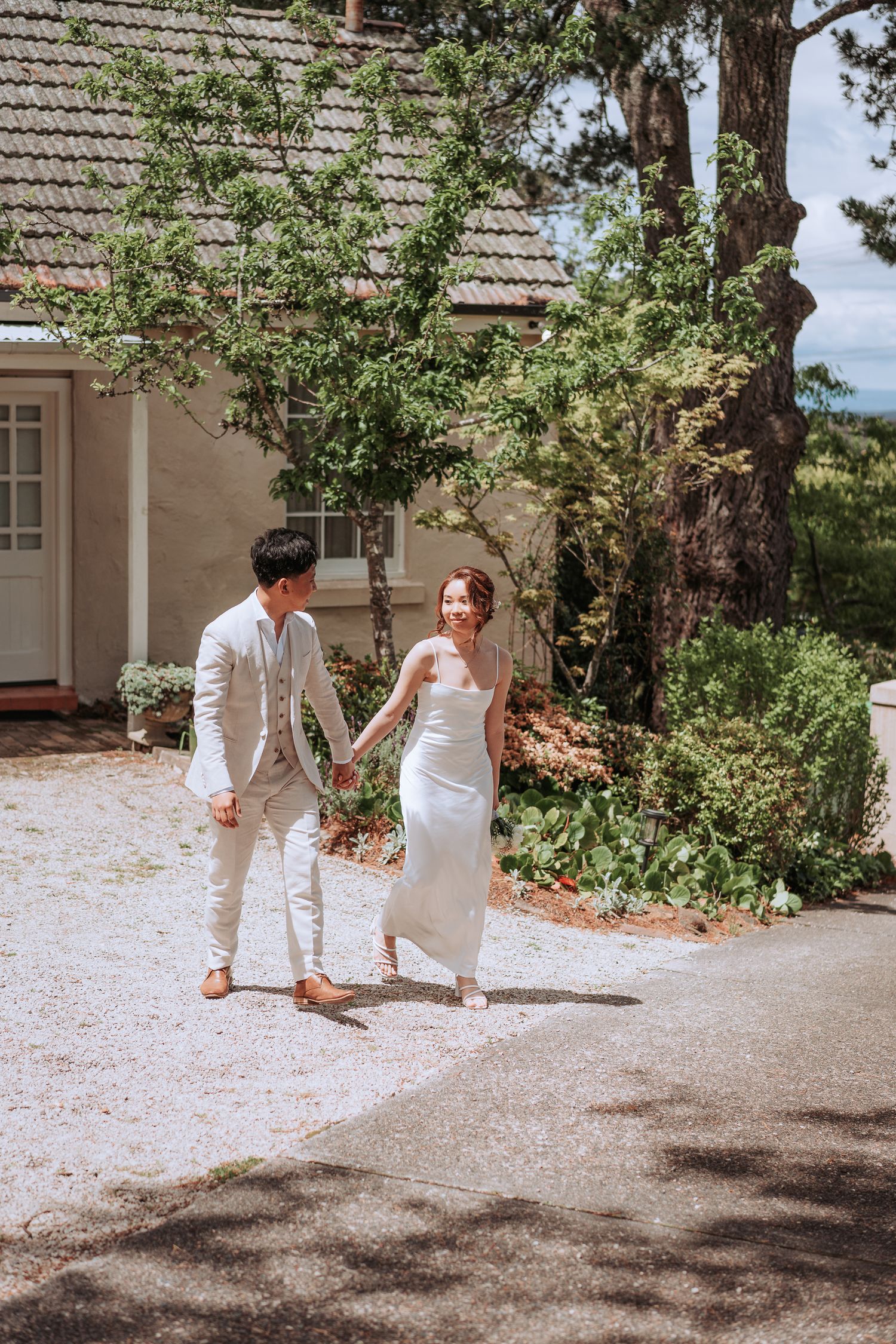 Couple in light-colored attire walking hand in hand on path by historic house.