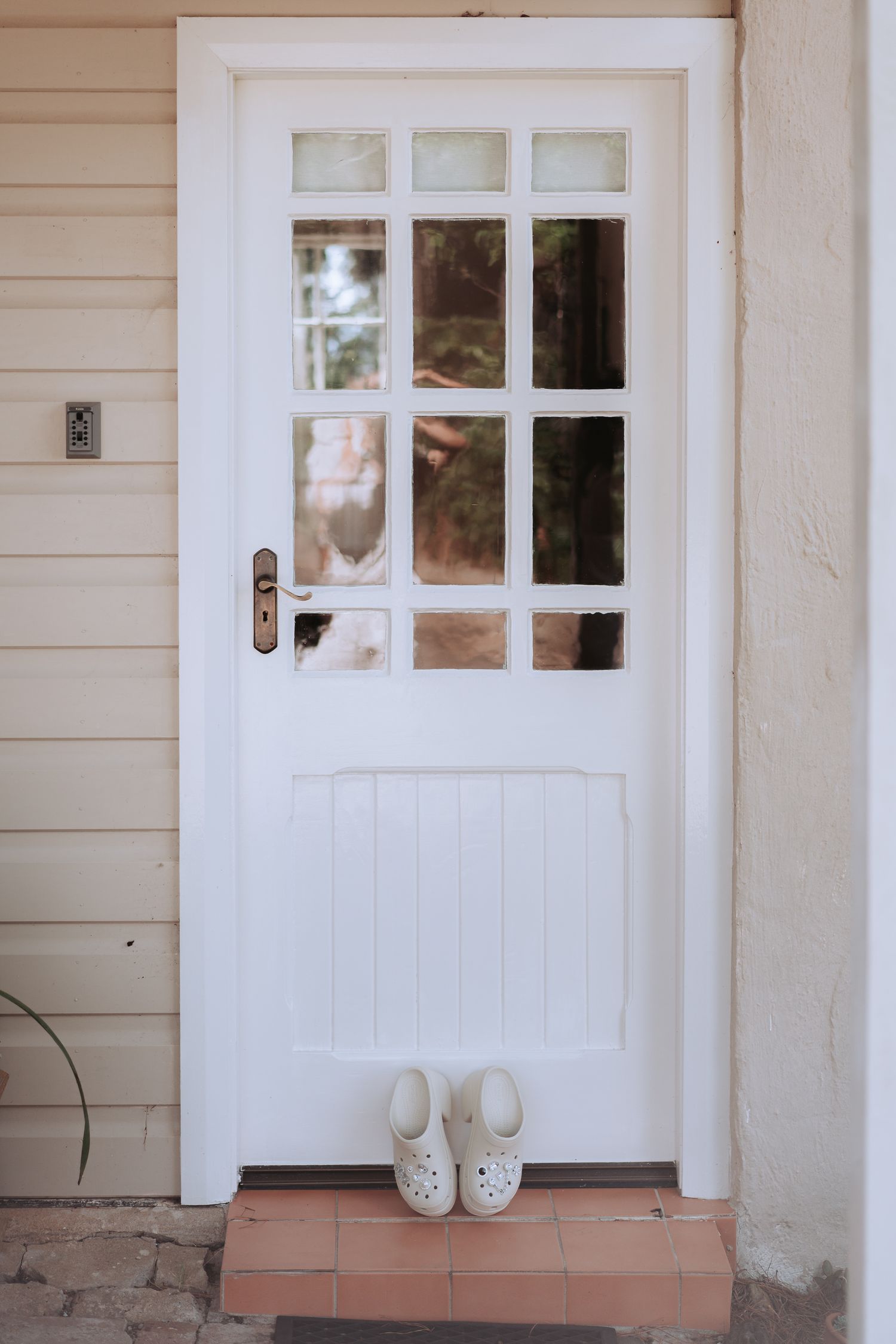 White door with glass panes and white clogs placed at doorstep of cream colored house.