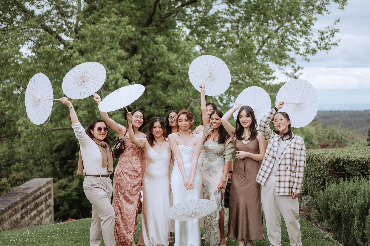 Wedding party holding white parasols outdoors with mountain vista in background.