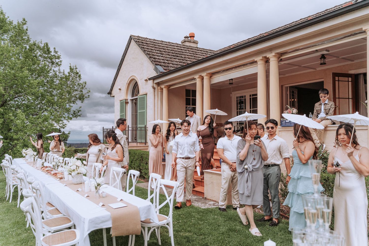 Elegant outdoor garden party with long white tables and guests in light summer attire gathered on a patio.