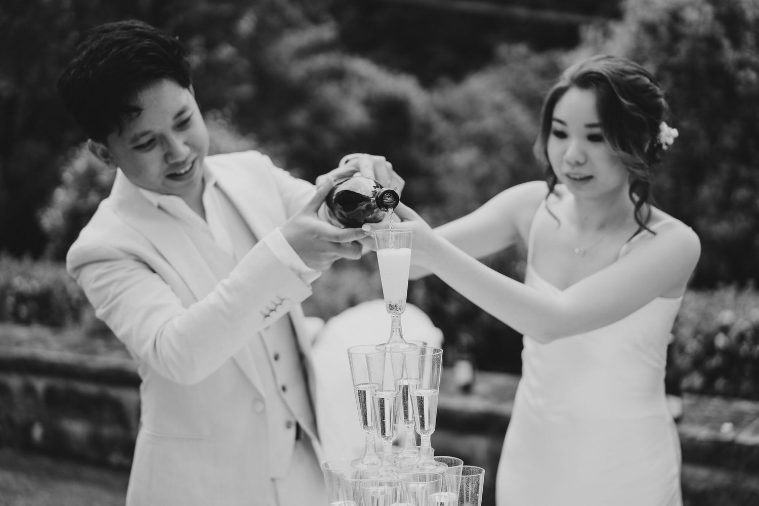 Black and white photo of couple pouring champagne together outdoors.
