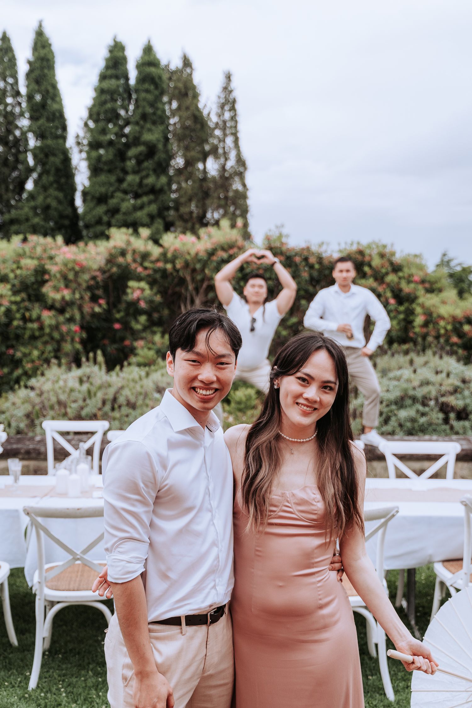 Guests in light colored outfits pose at an outdoor garden event with tall cypress trees in background.