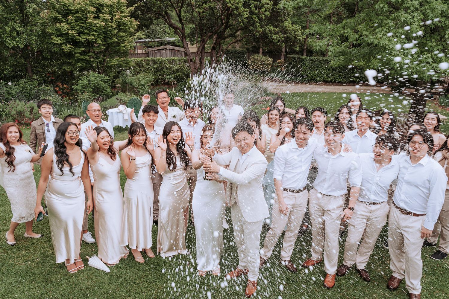 Large group in all-white attire celebrates with champagne spray in a garden setting.