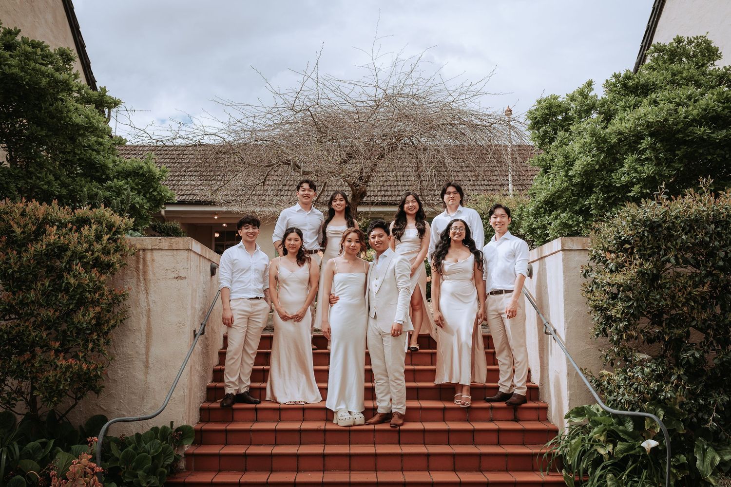 Wedding party in white attire poses on outdoor stairs with decorative landscaping.