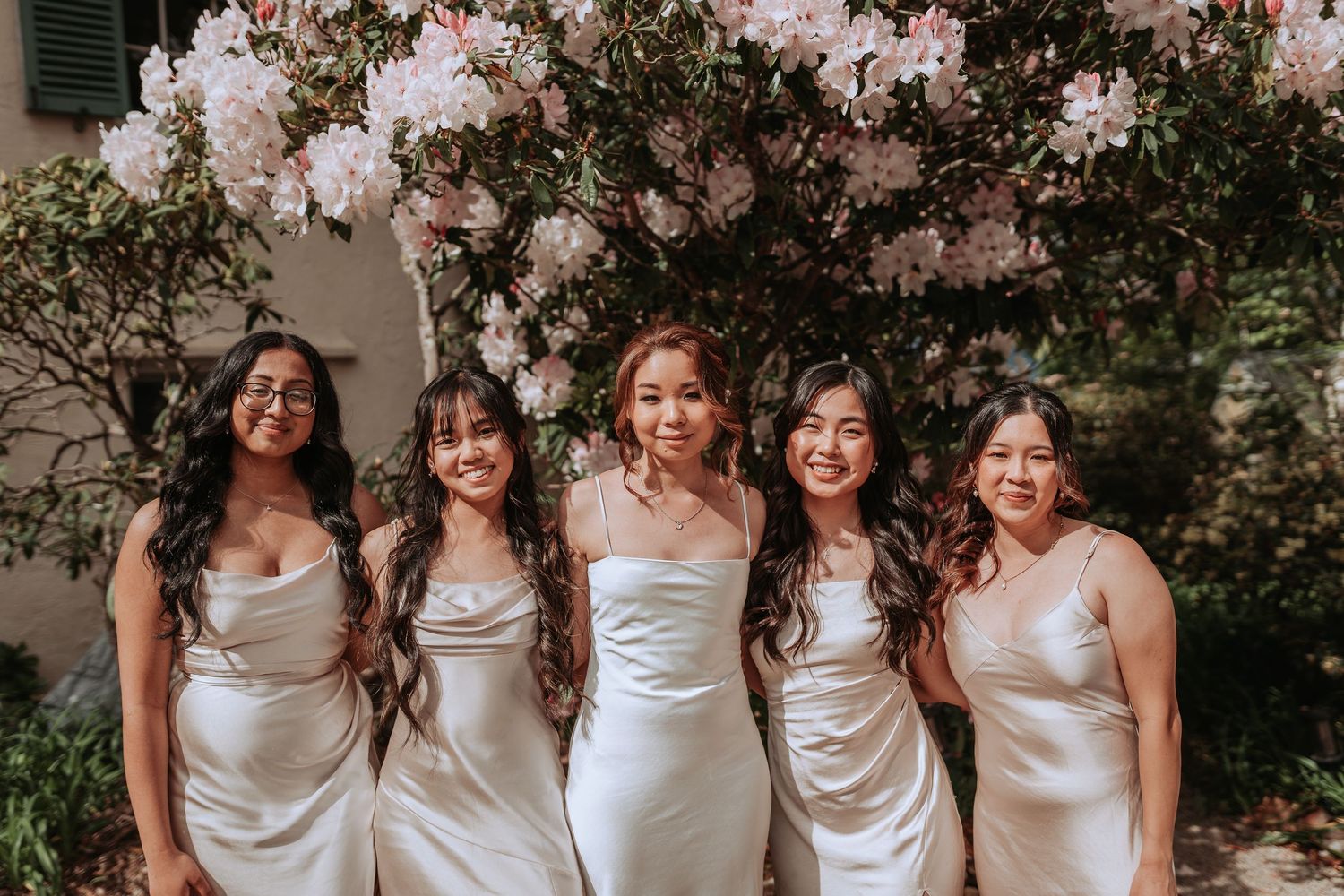 Bridesmaids in silver satin dresses pose together in front of blooming flowers.