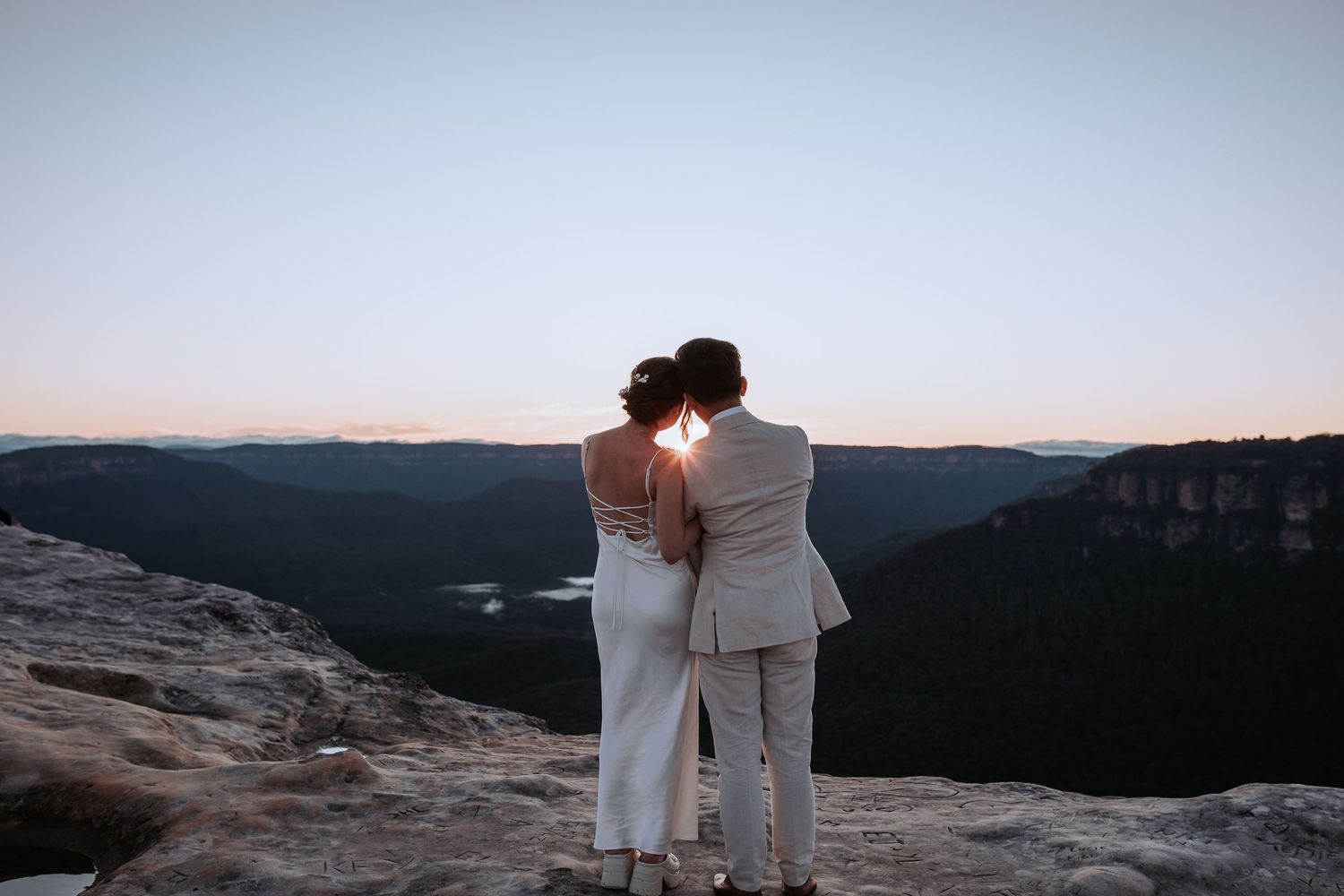 Couple embraces while overlooking mountain vista at sunset.