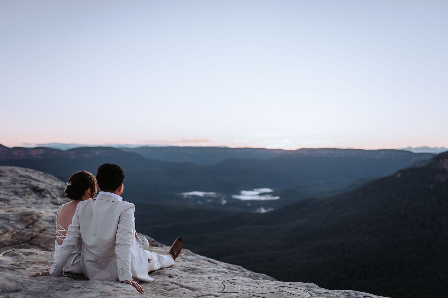A couple sits together on a rocky cliff edge overlooking a misty mountain valley at dusk.
