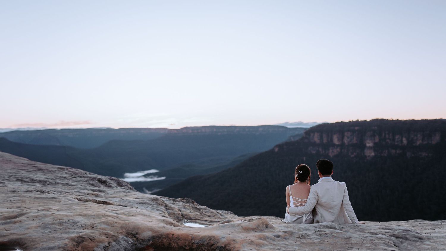 Two people perched on a cliff edge admiring scenic mountain views across a deep valley.