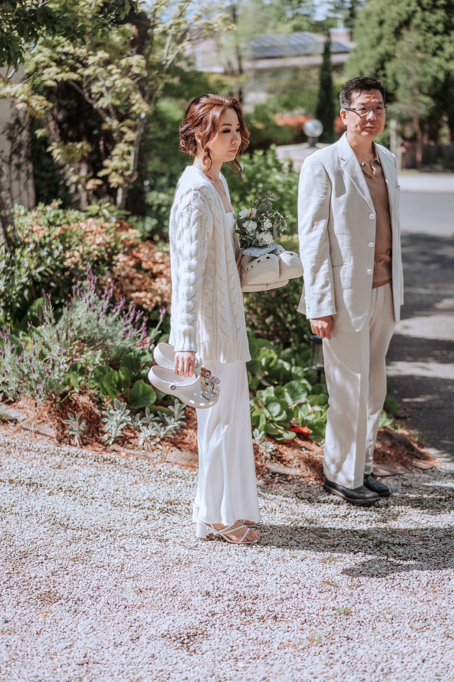 A couple in matching white outfits stands on a gravel path surrounded by lush garden landscaping.