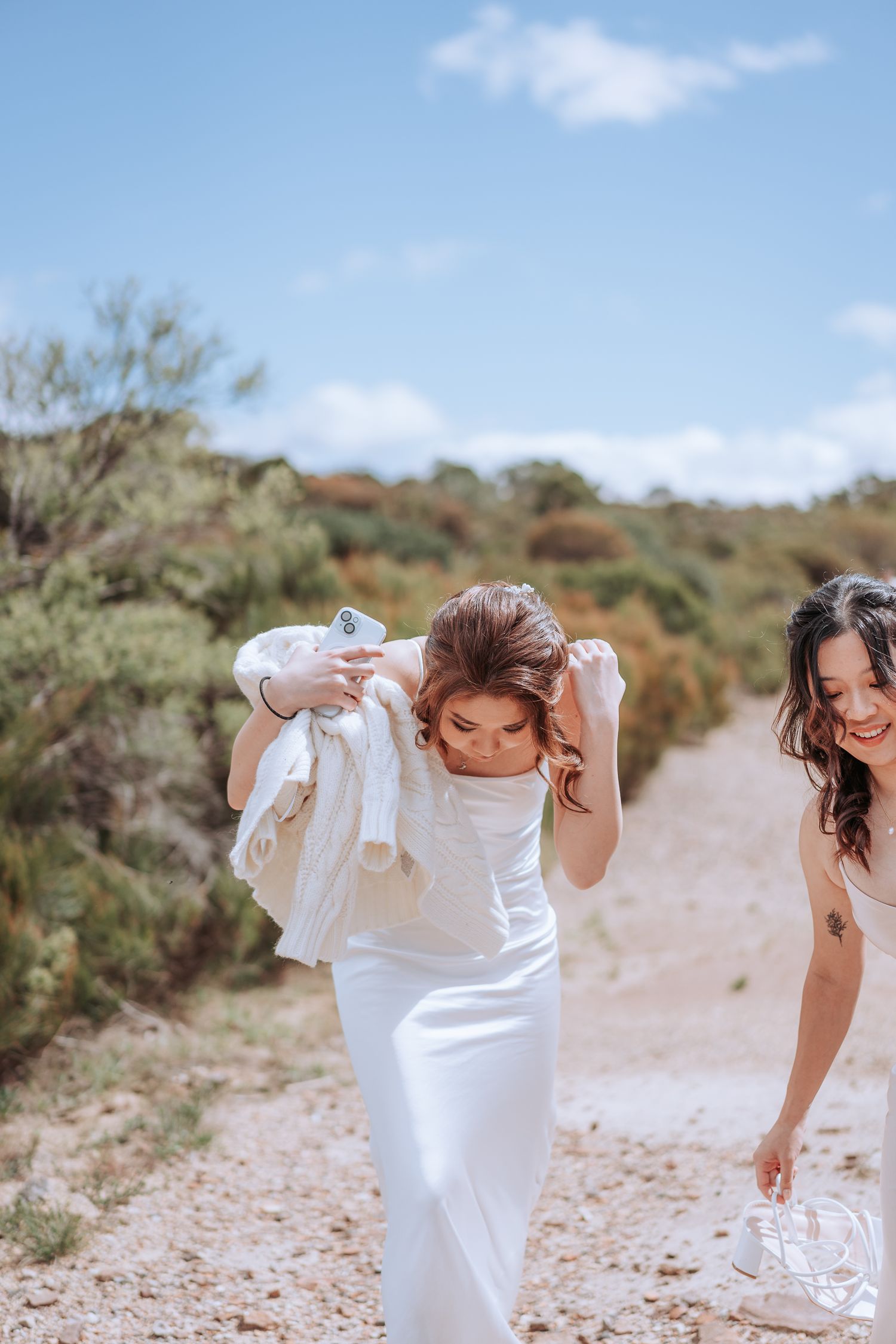 Two people in white attire walking along a sandy trail with natural bush surroundings.