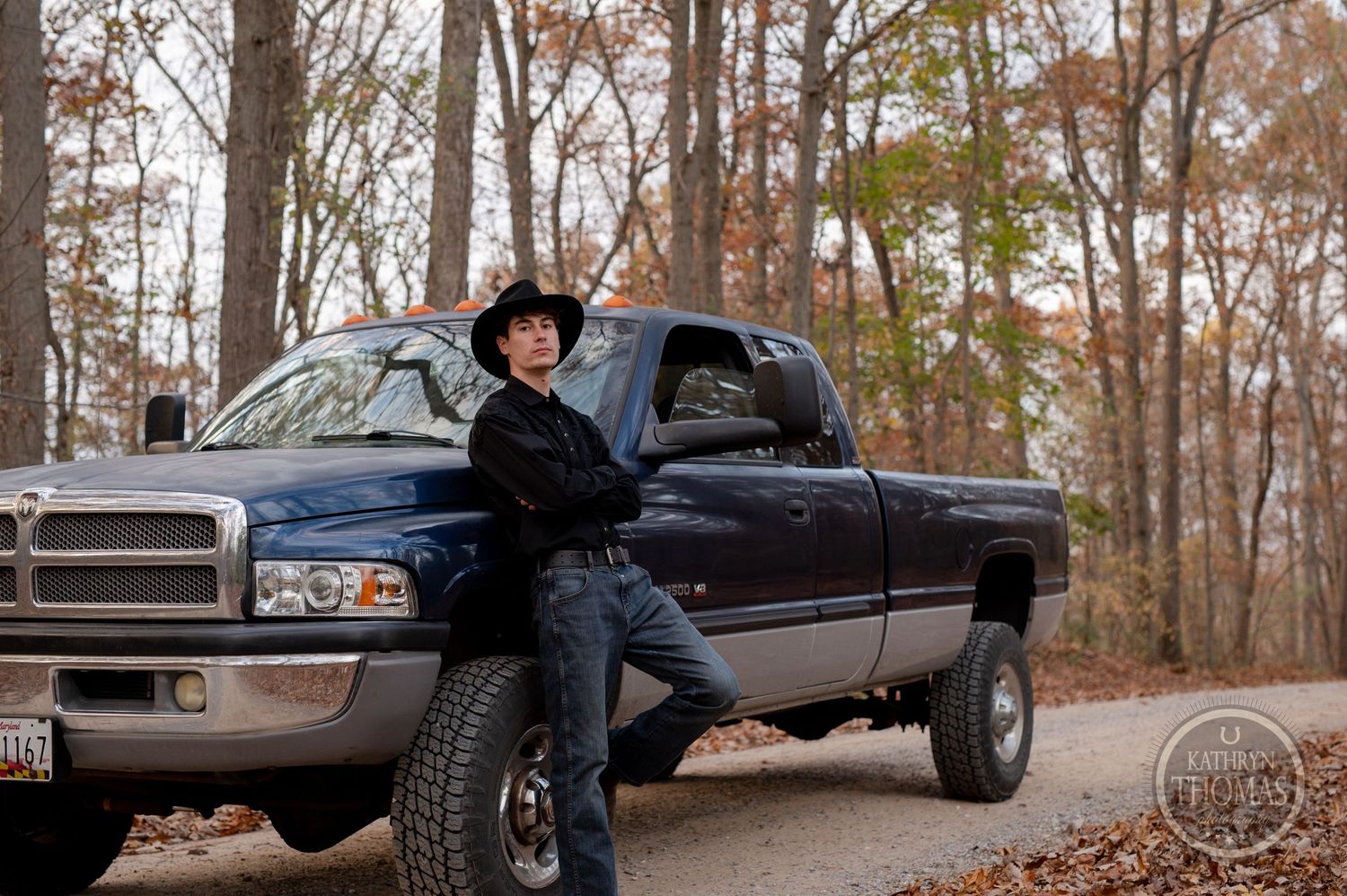 A pickup truck with someone in western wear leaning against it in a wooded autumn setting.