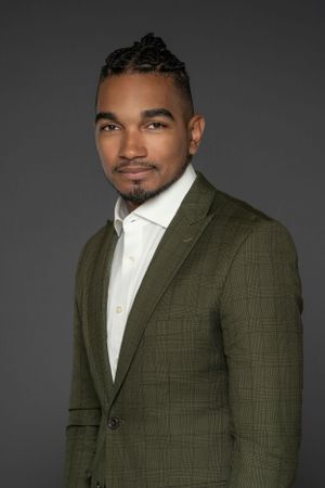 Professional man with braided hair wearing a green plaid blazer and white shirt against gray background.
