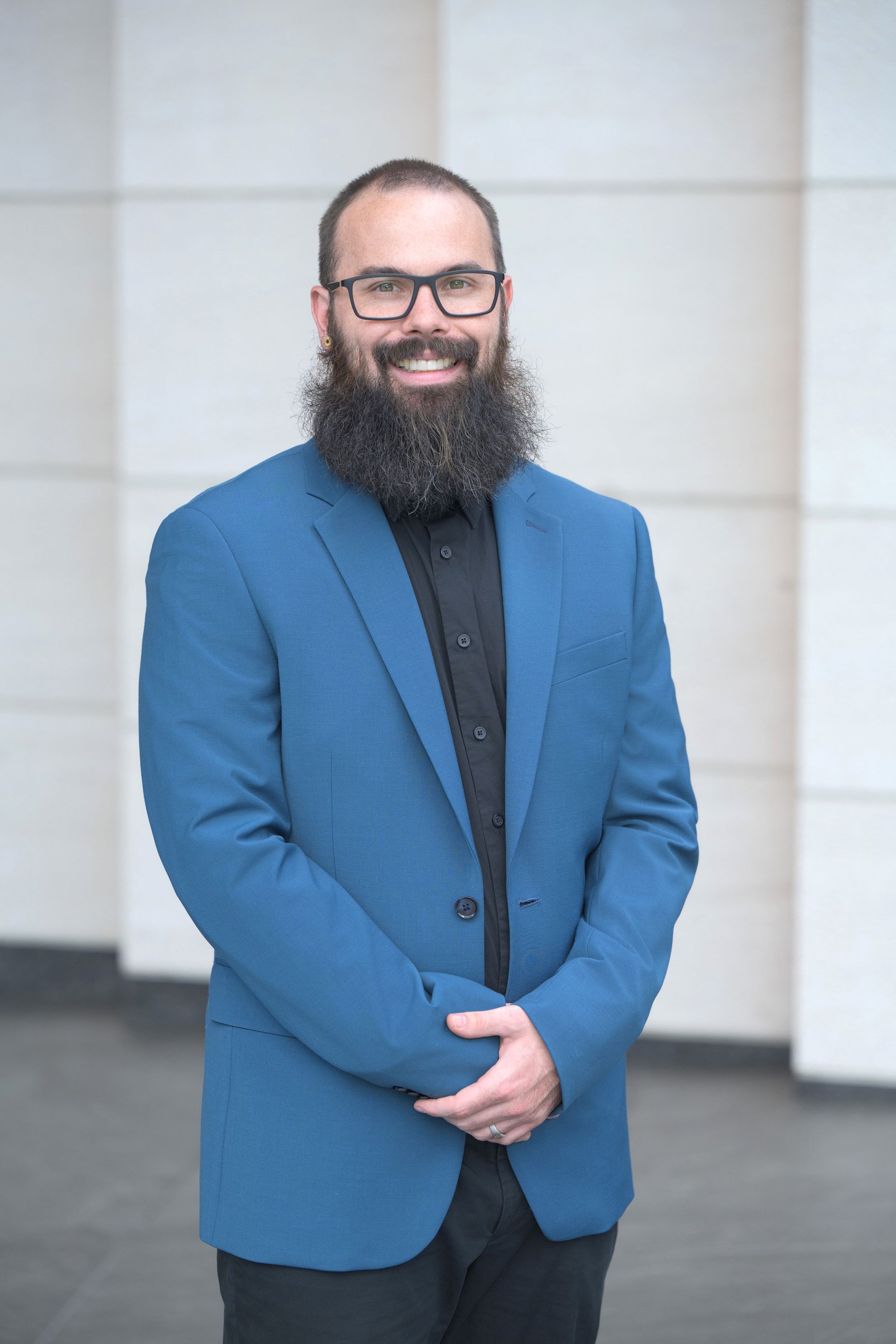 Smiling bearded man with glasses wearing a blue blazer and black shirt in a professional headshot.