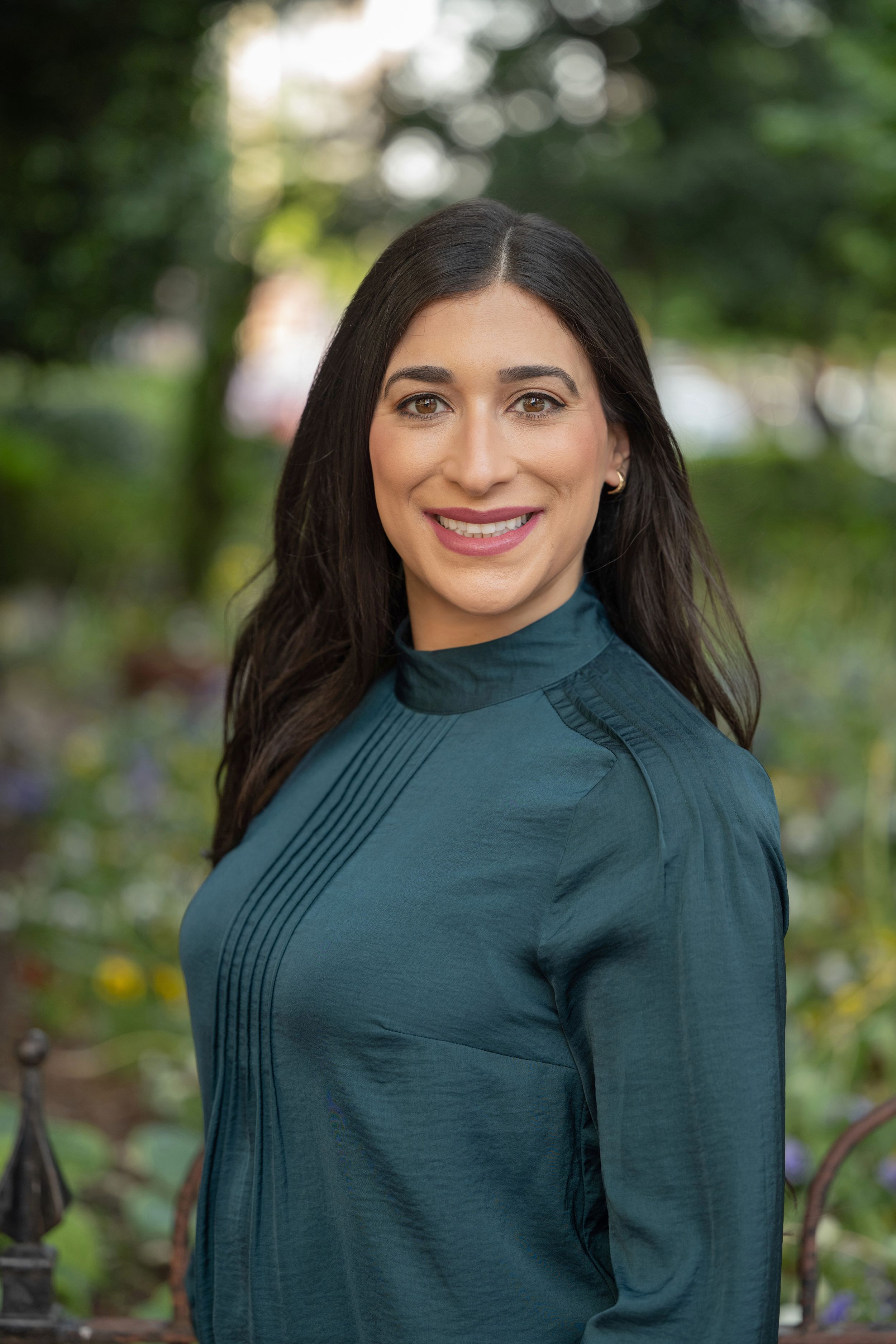 Smiling woman with long dark hair wearing a teal blouse in an outdoor professional portrait.