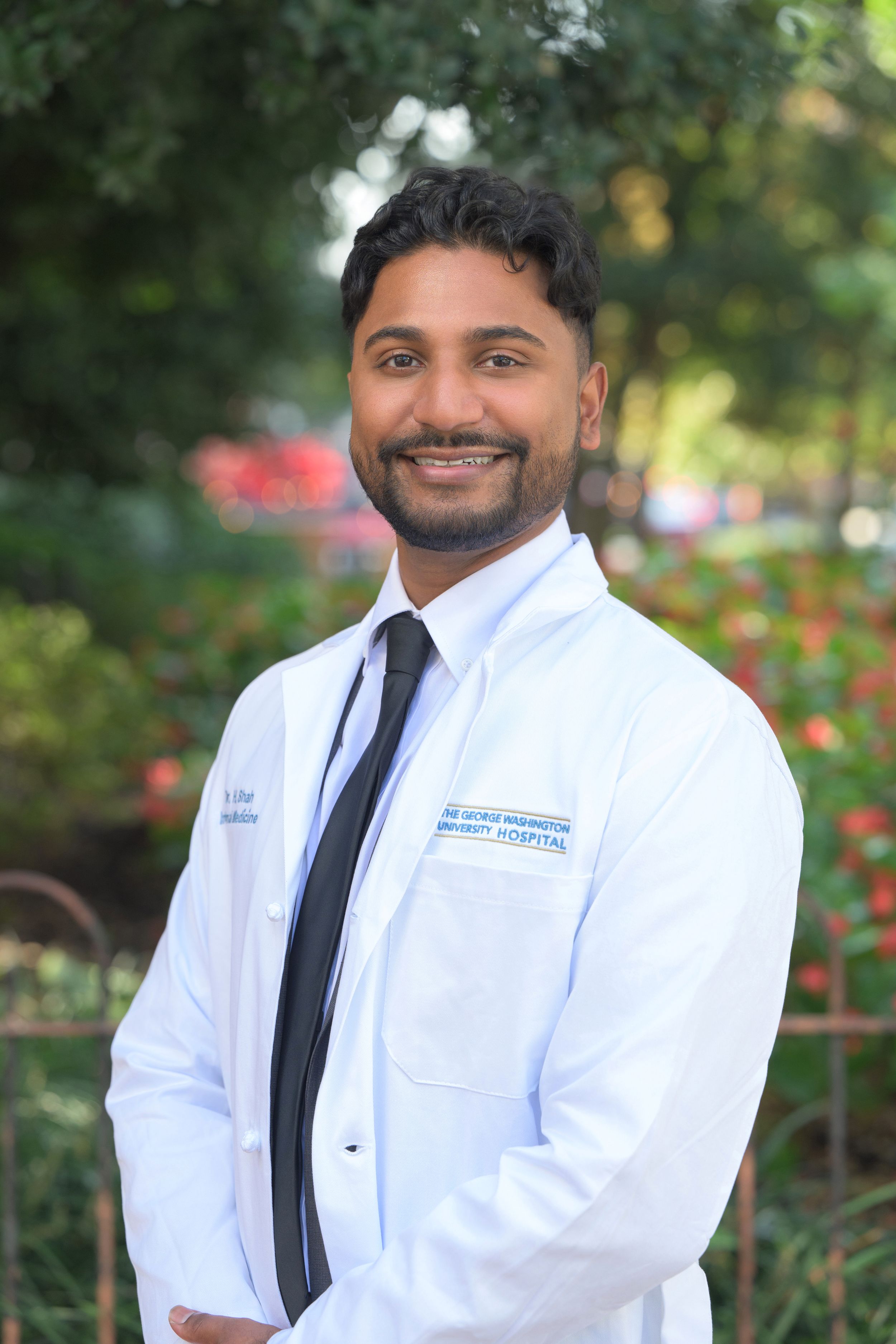 Young male doctor smiling in a white medical coat with a black tie in an outdoor portrait.