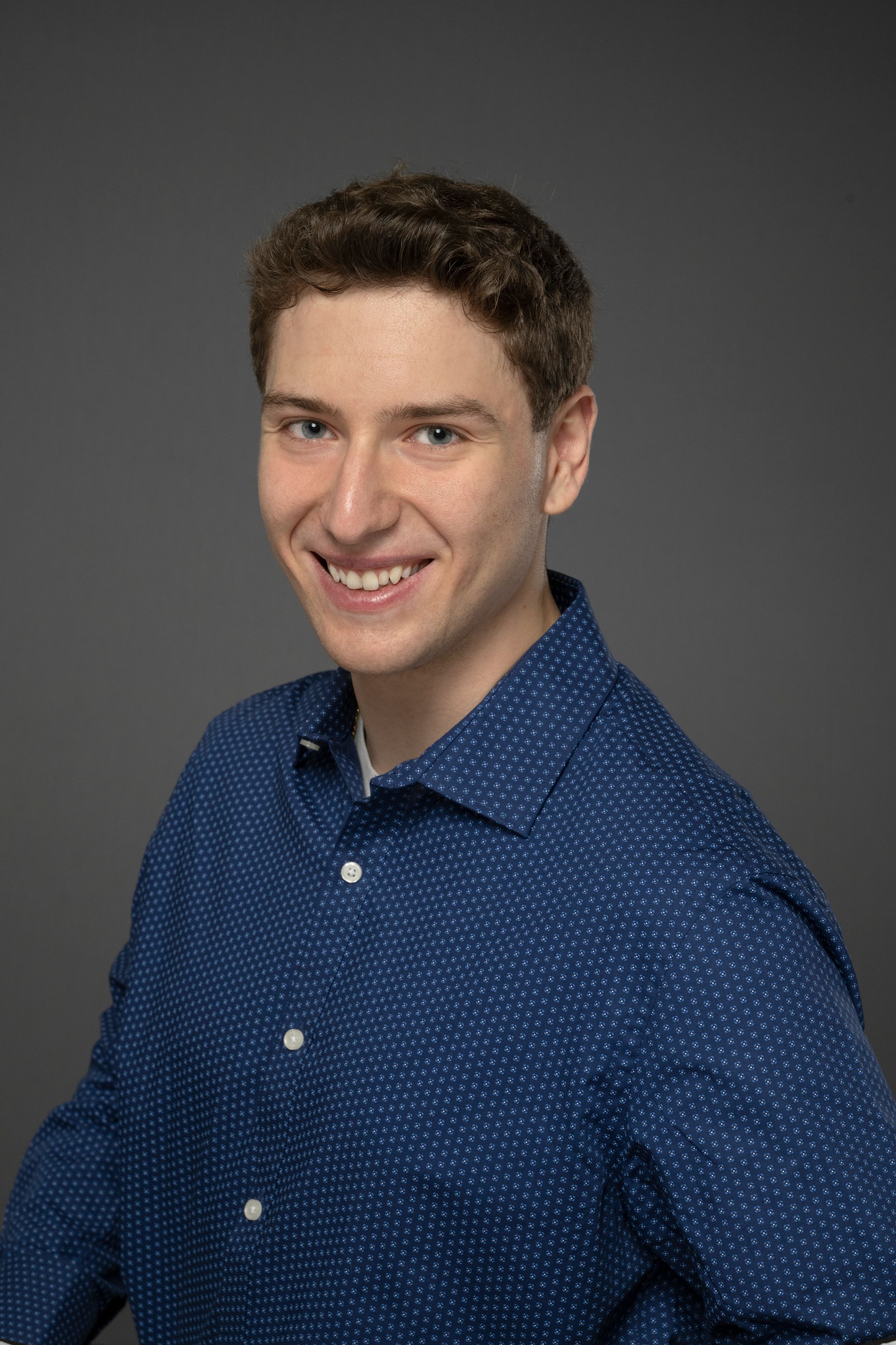 Young smiling man in a blue patterned shirt poses for a professional headshot on gray background.