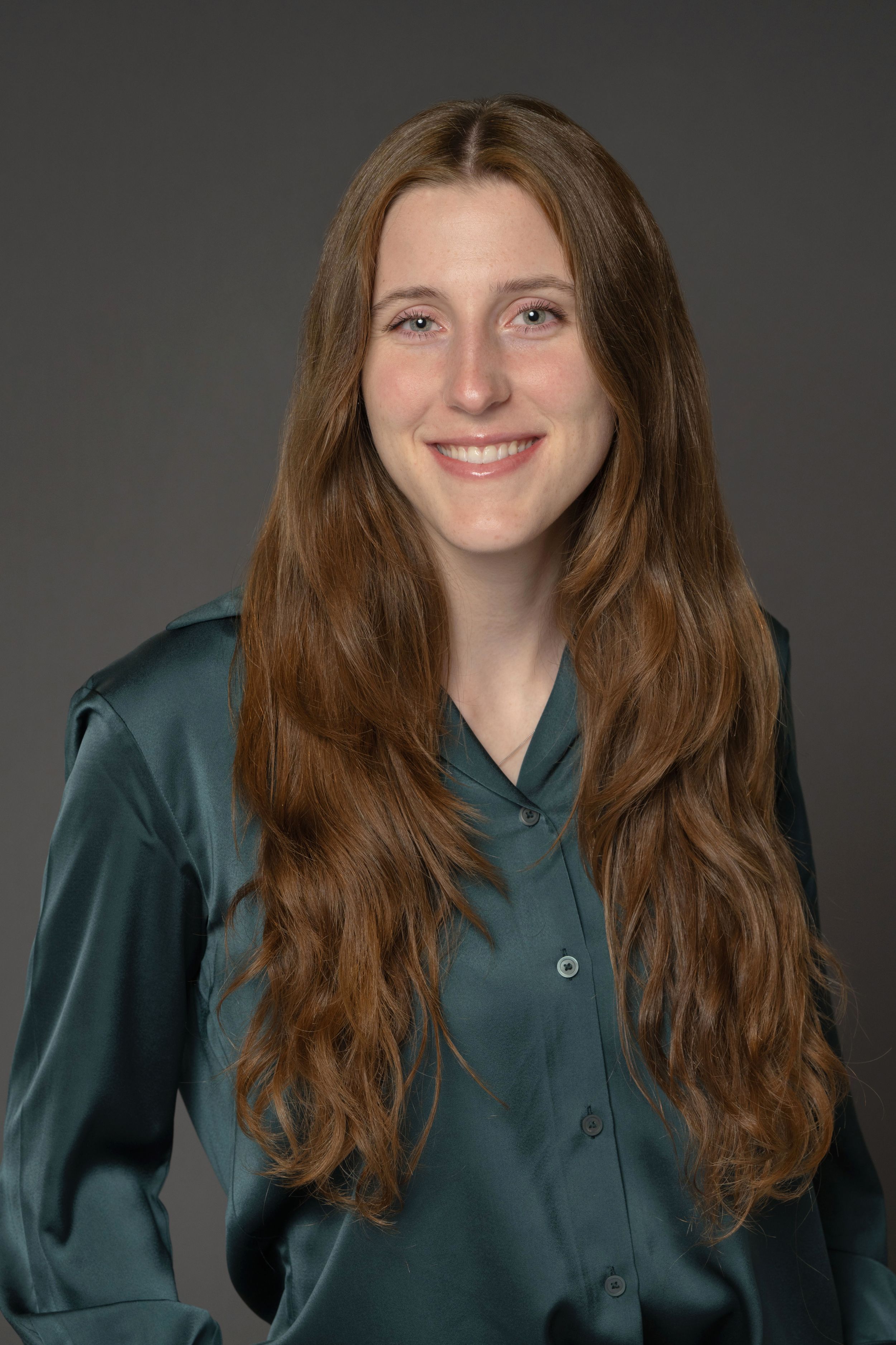 Young woman with long auburn hair smiling, wearing a teal satin blouse against a gray background.