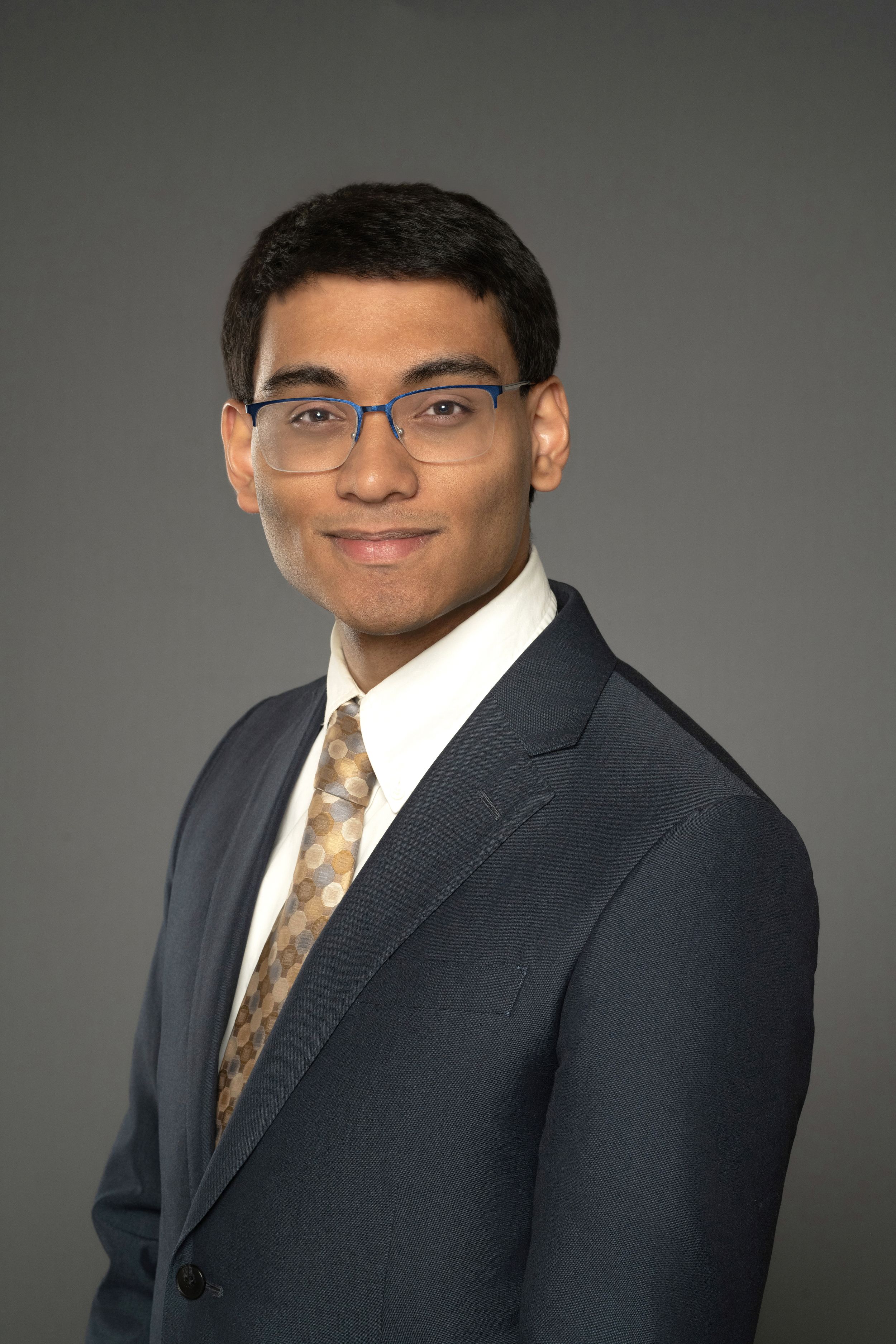 Young man wearing glasses, a navy suit, and patterned tie in a formal professional headshot.