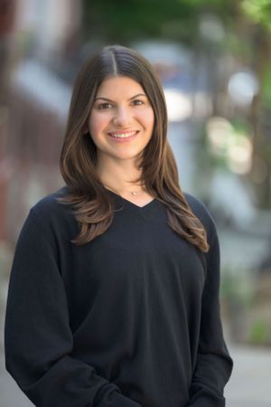 Young woman with long brown hair smiling outdoors in a black v-neck sweater with blurred street background.