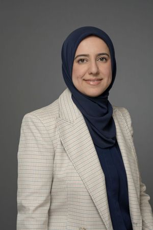 Professional woman wearing a navy hijab and plaid blazer smiling against a gray background.