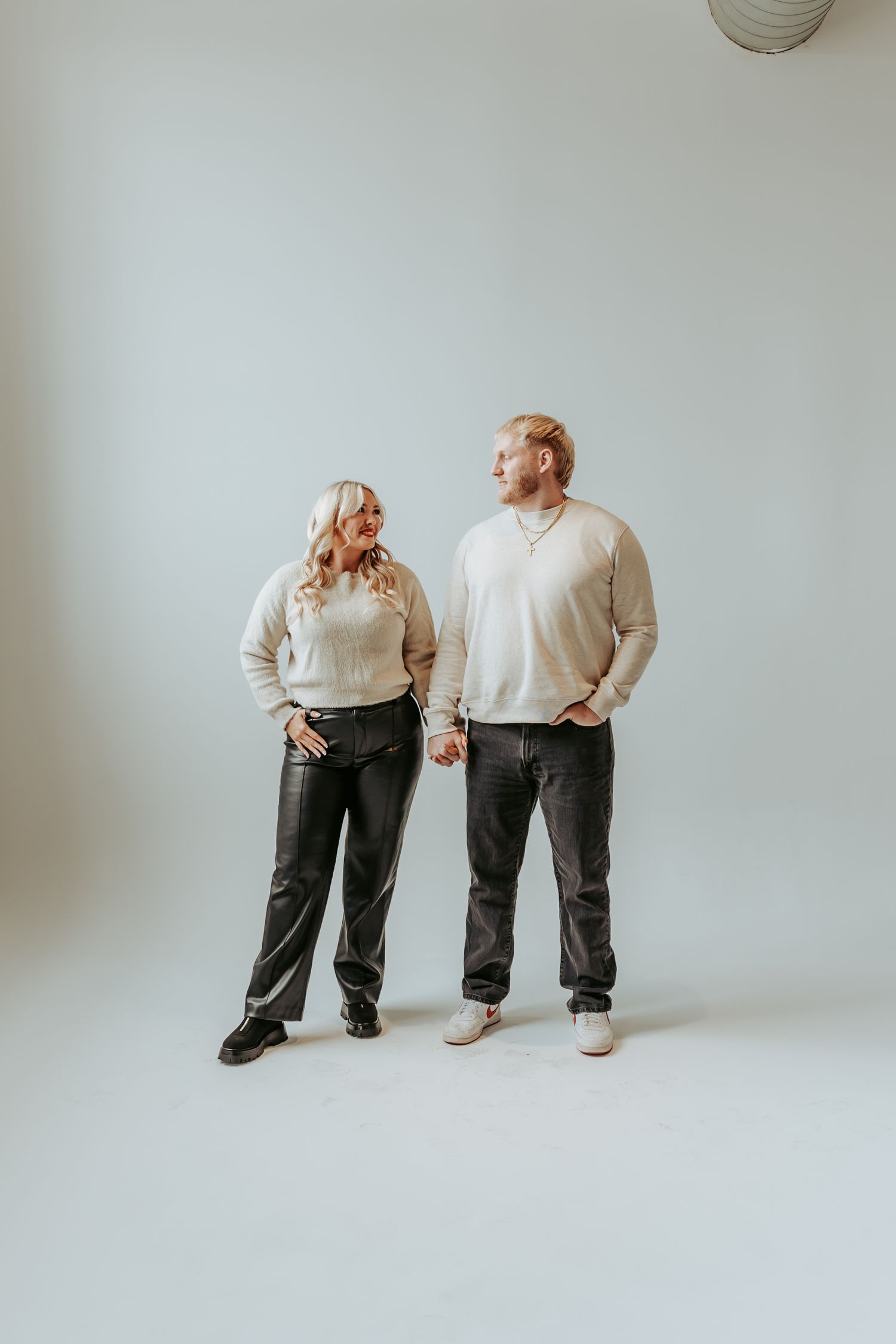 Couple in cream sweaters and dark jeans standing casually against white background.