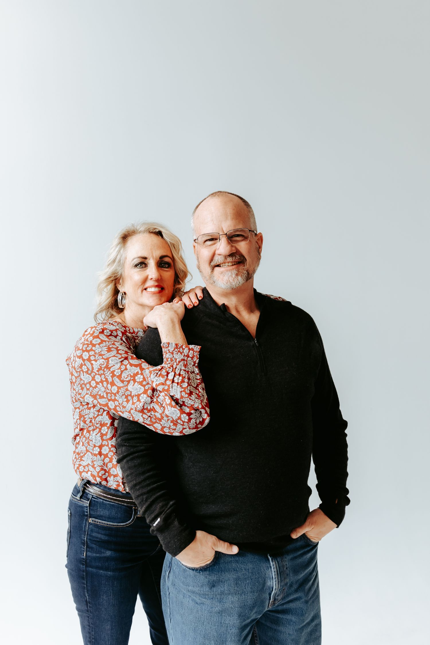 Couple posing against white wall, woman in floral top and man in black sweater.