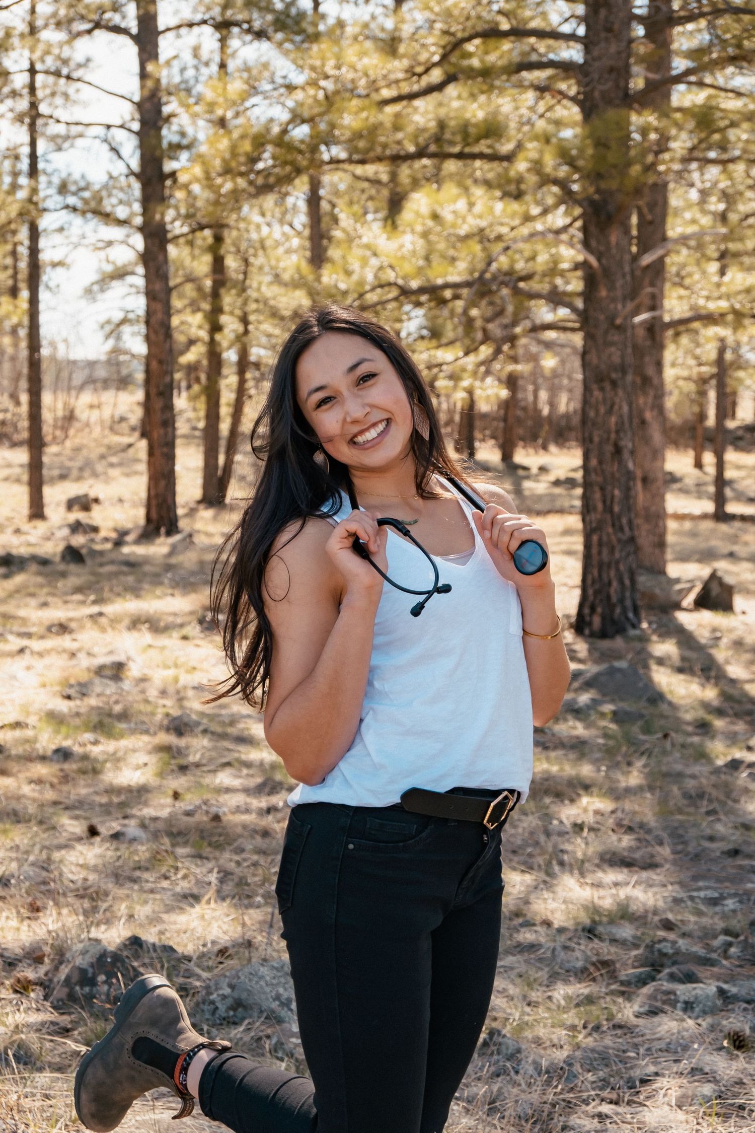 A healthcare professional poses outdoors in workout attire with a stethoscope against a backdrop of pine trees in sunlight.