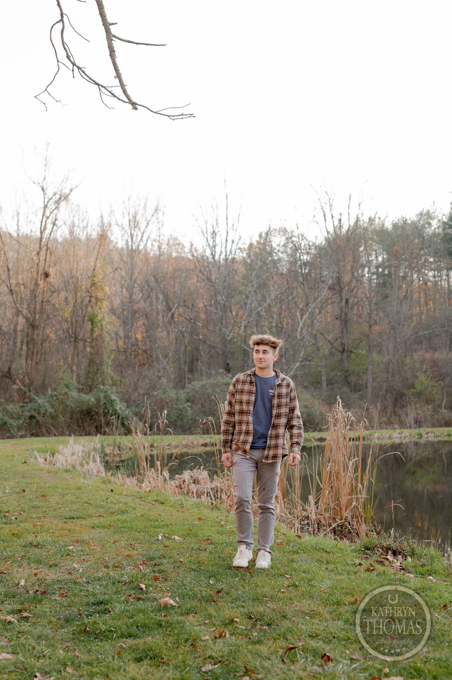 A person in plaid and jeans stands on grassy lakeside with bare trees in background.