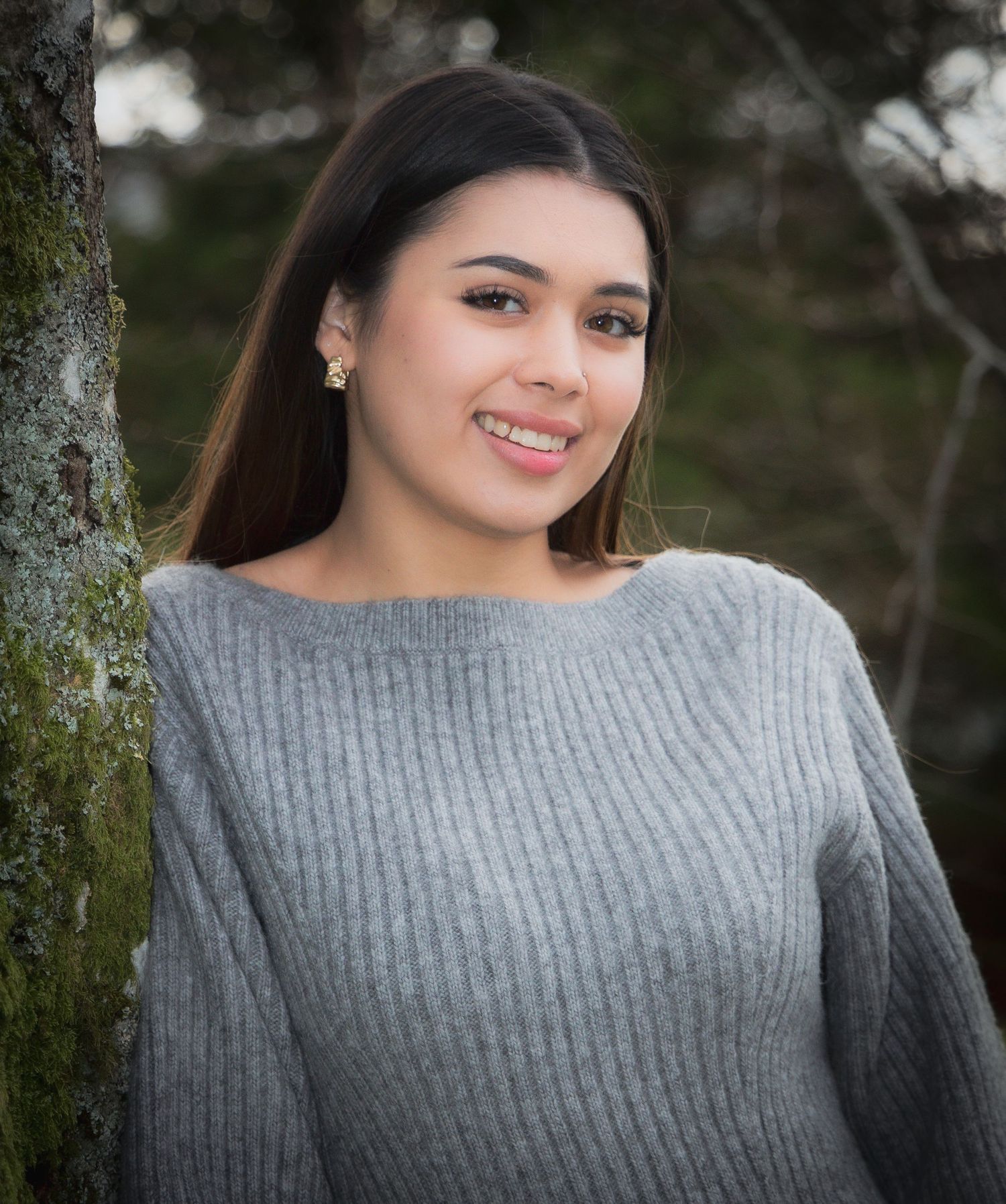 Young woman with long dark hair smiling outdoors in a grey sweater beside a tree.