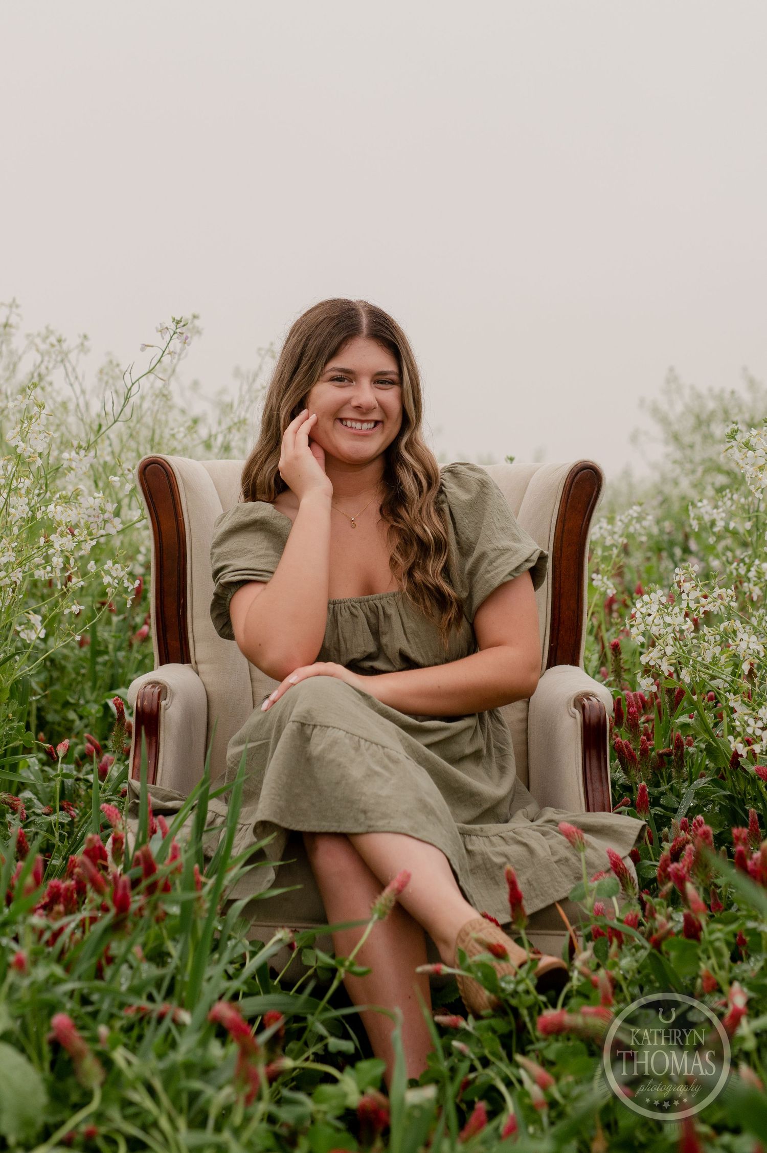 A rustic outdoor portrait featuring someone in an olive dress sitting in a brown armchair surrounded by wildflowers.