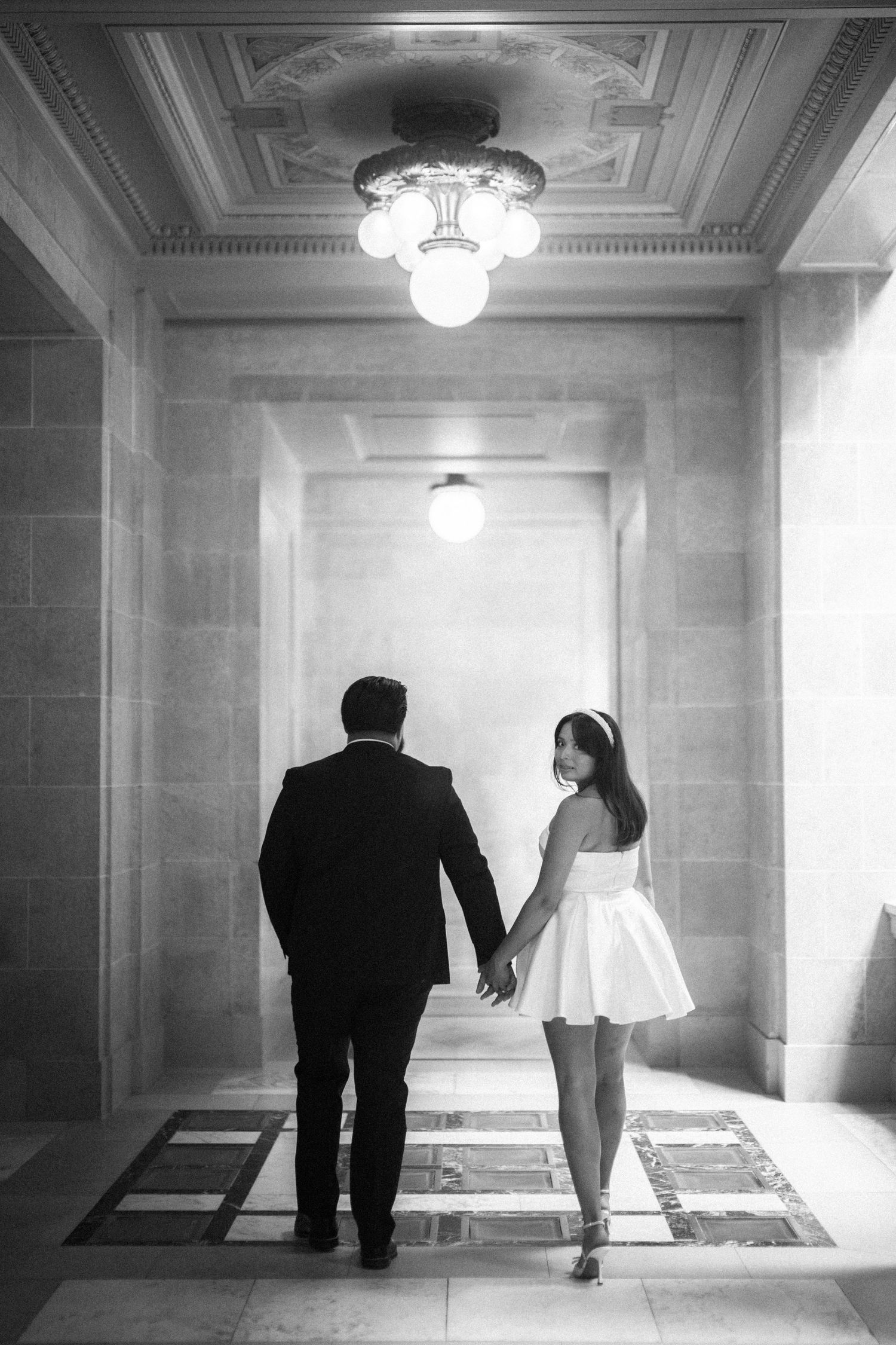 A couple holding hands walking down an elegant hallway with marble floors and ornate lighting fixtures.