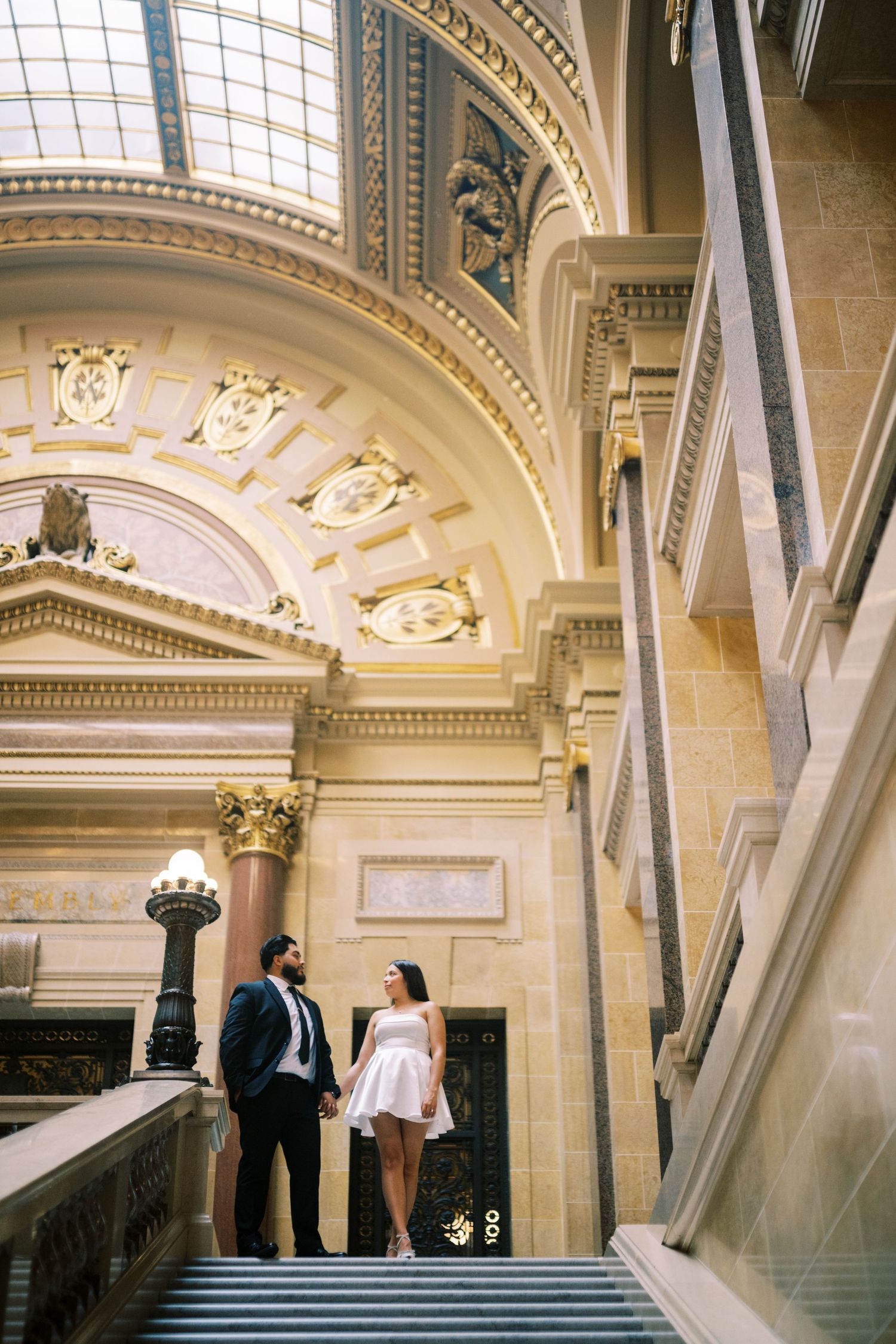 A couple stands on an ornate staircase inside a grand historic building with neoclassical architectural details.