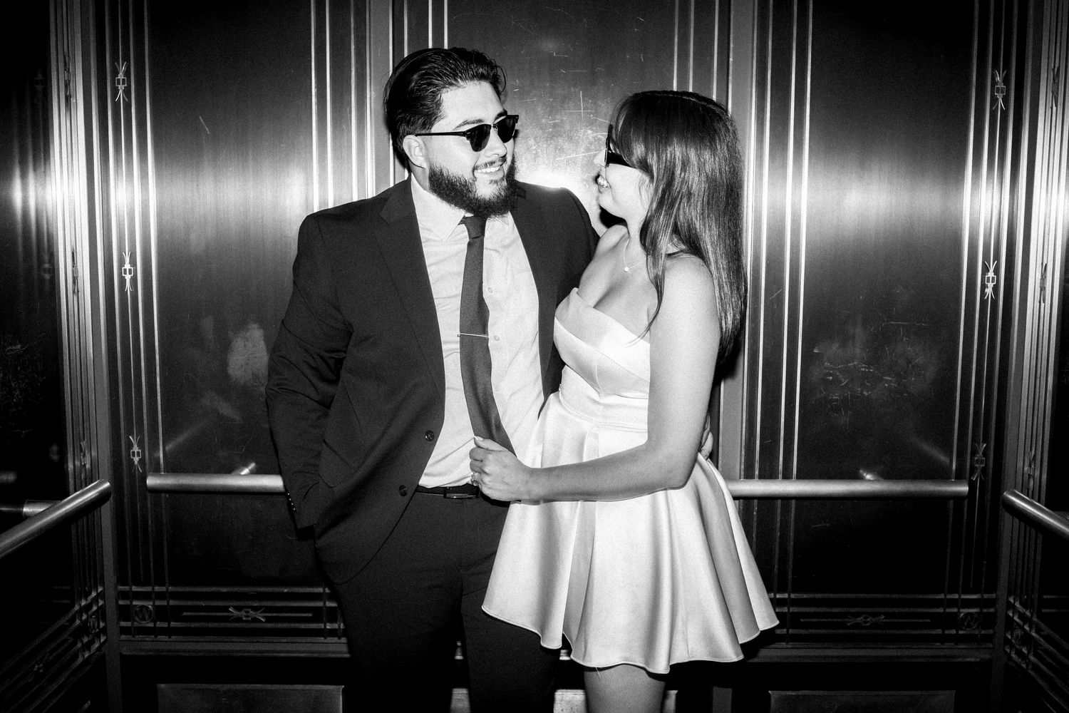 A stylish couple in black and white embraces in an elevator, with him in a suit and her in a white dress.