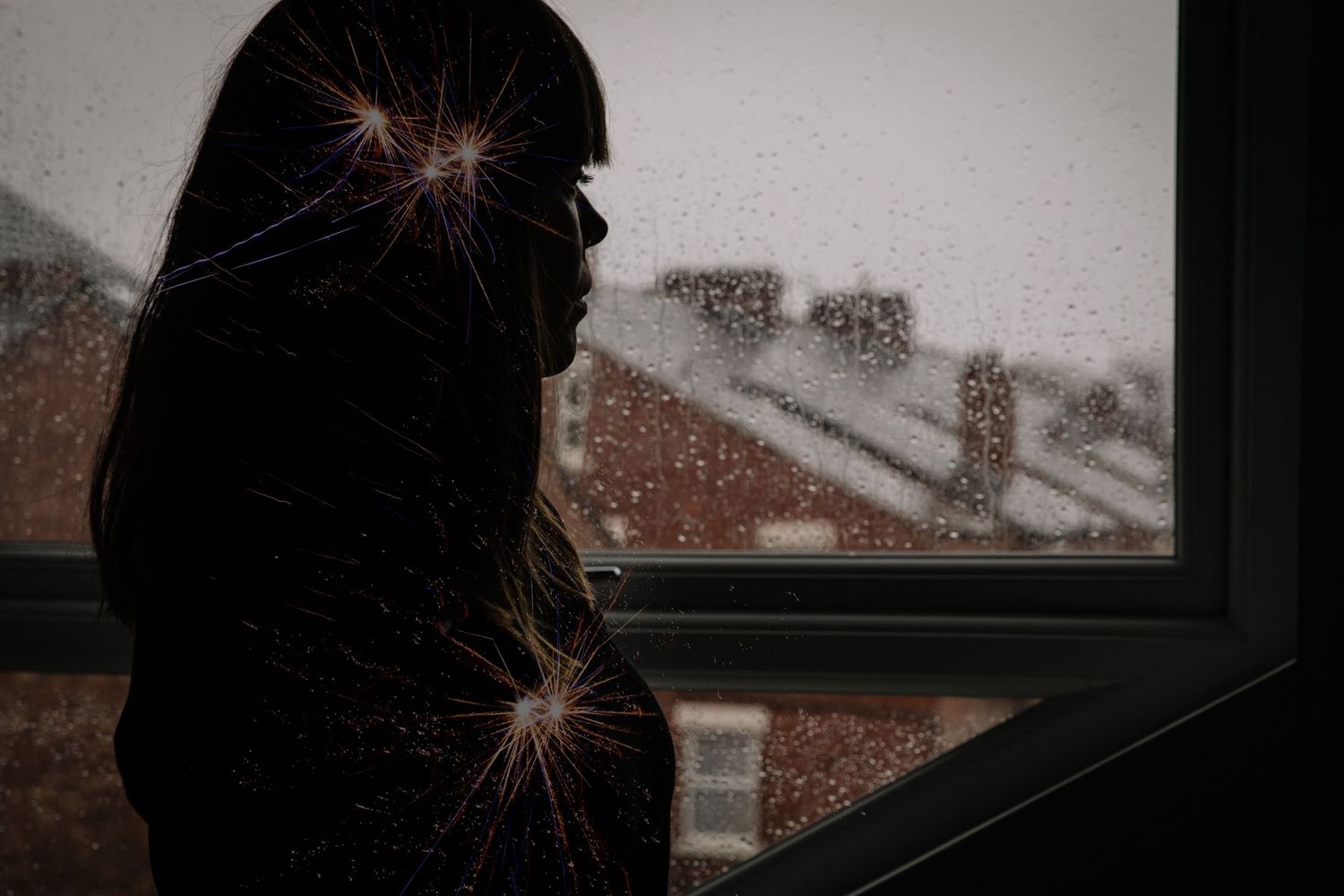 A silhouetted figure gazes out a window on a snowy winter evening as lens flares sparkle across the dark scene.