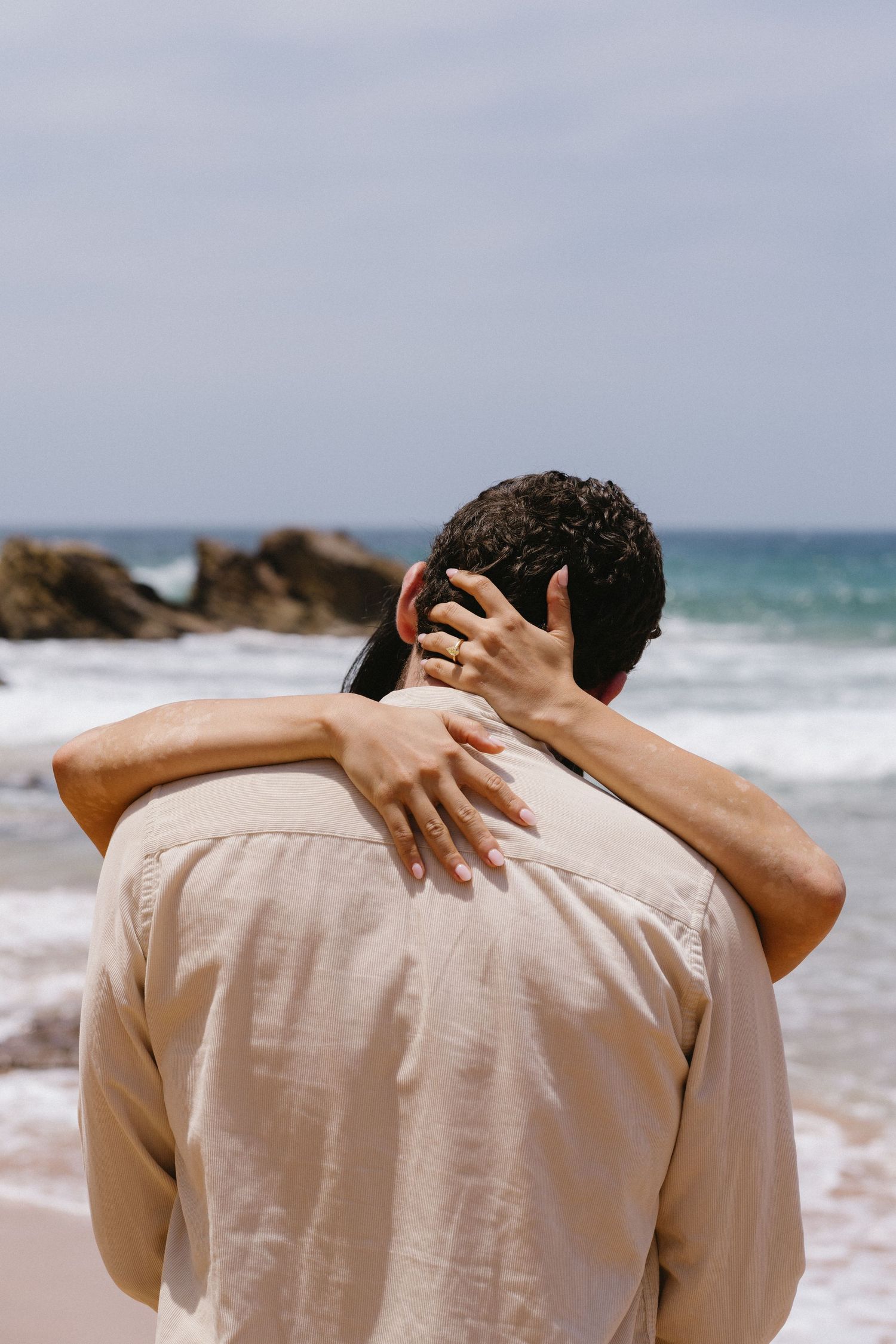 A close-up back view of an embrace on a rocky beach with waves in the background.